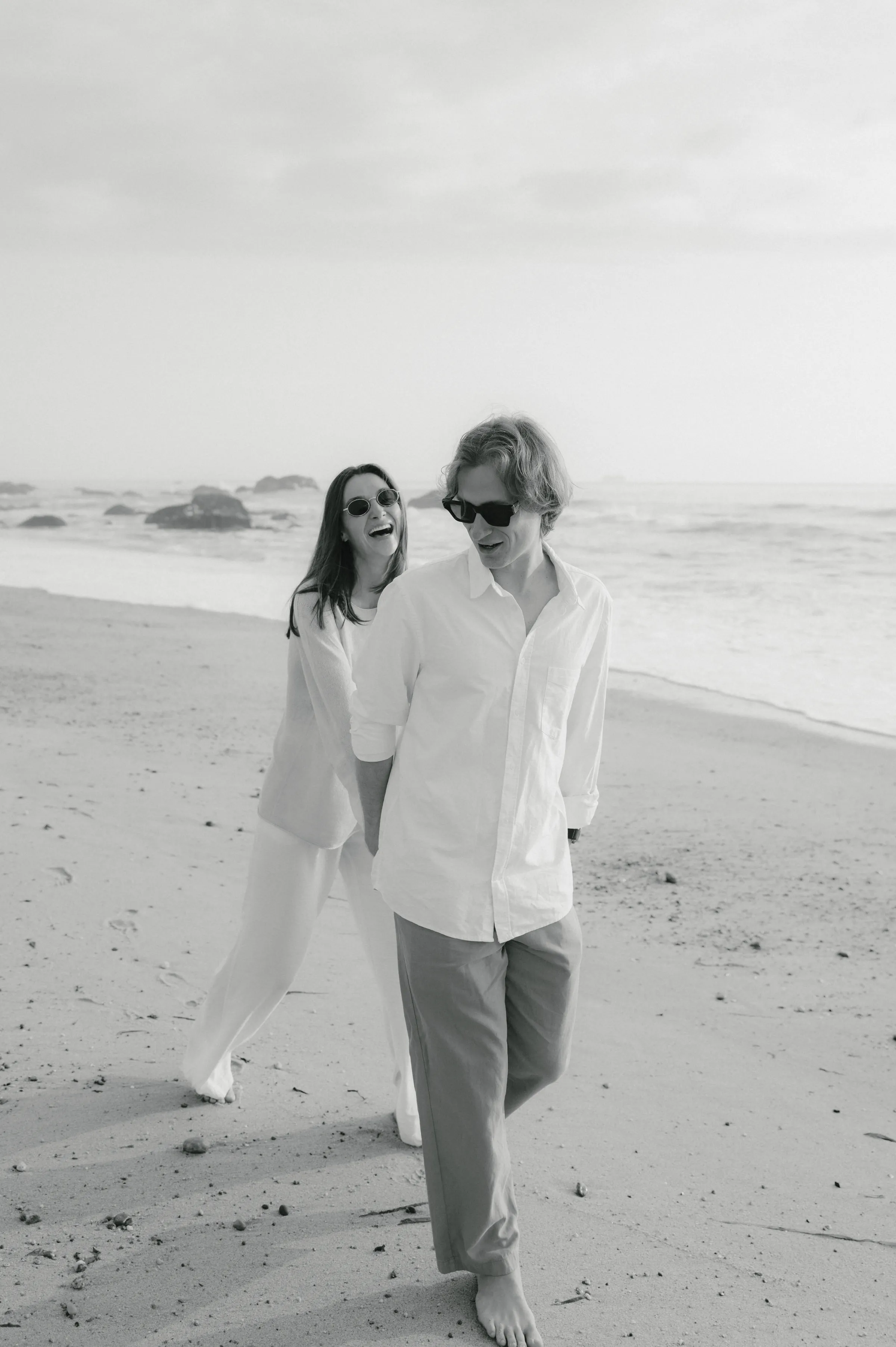Couple walking barefoot on a sandy beach during a black-and-white seaside session.