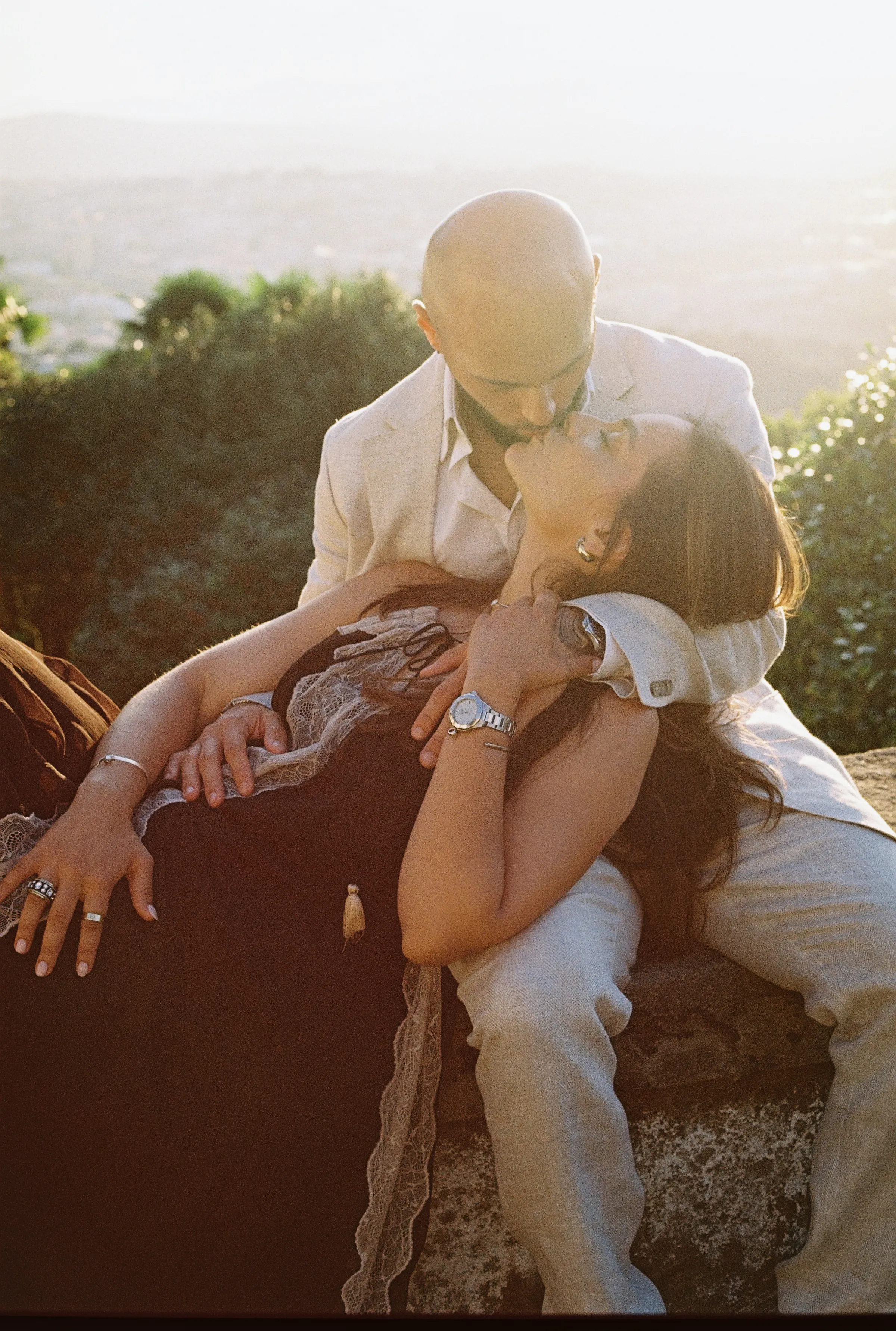 Couple seated close during an outdoor session in soft light, with greenery and distant hills behind them.