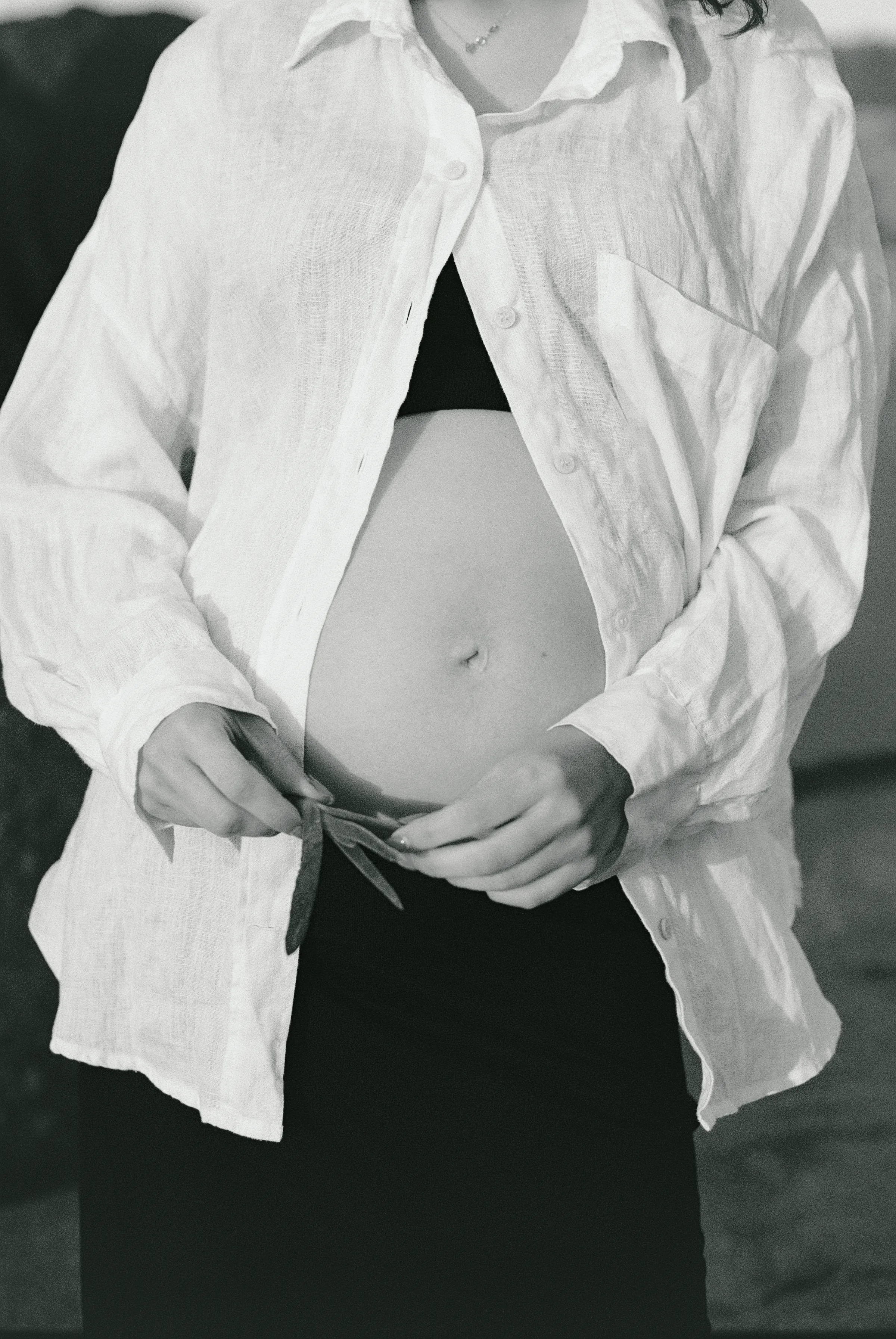 Black-and-white maternity close portrait with hands framing a pregnant belly under an open white shirt.