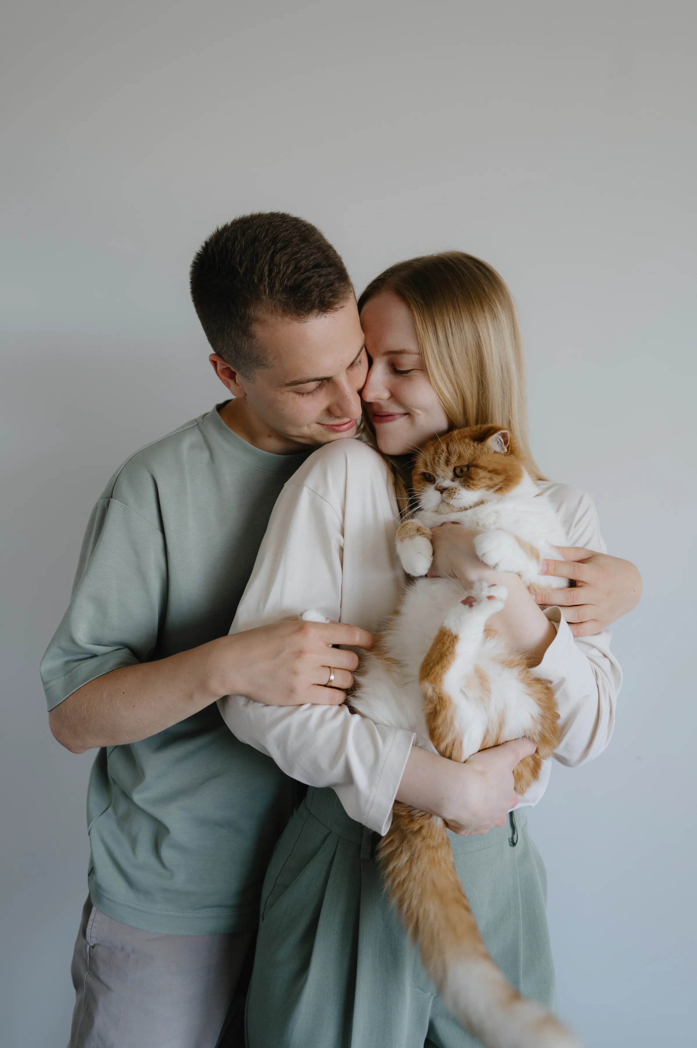 Indoor close portrait of a couple holding an orange-and-white cat against a plain wall in soft light.
