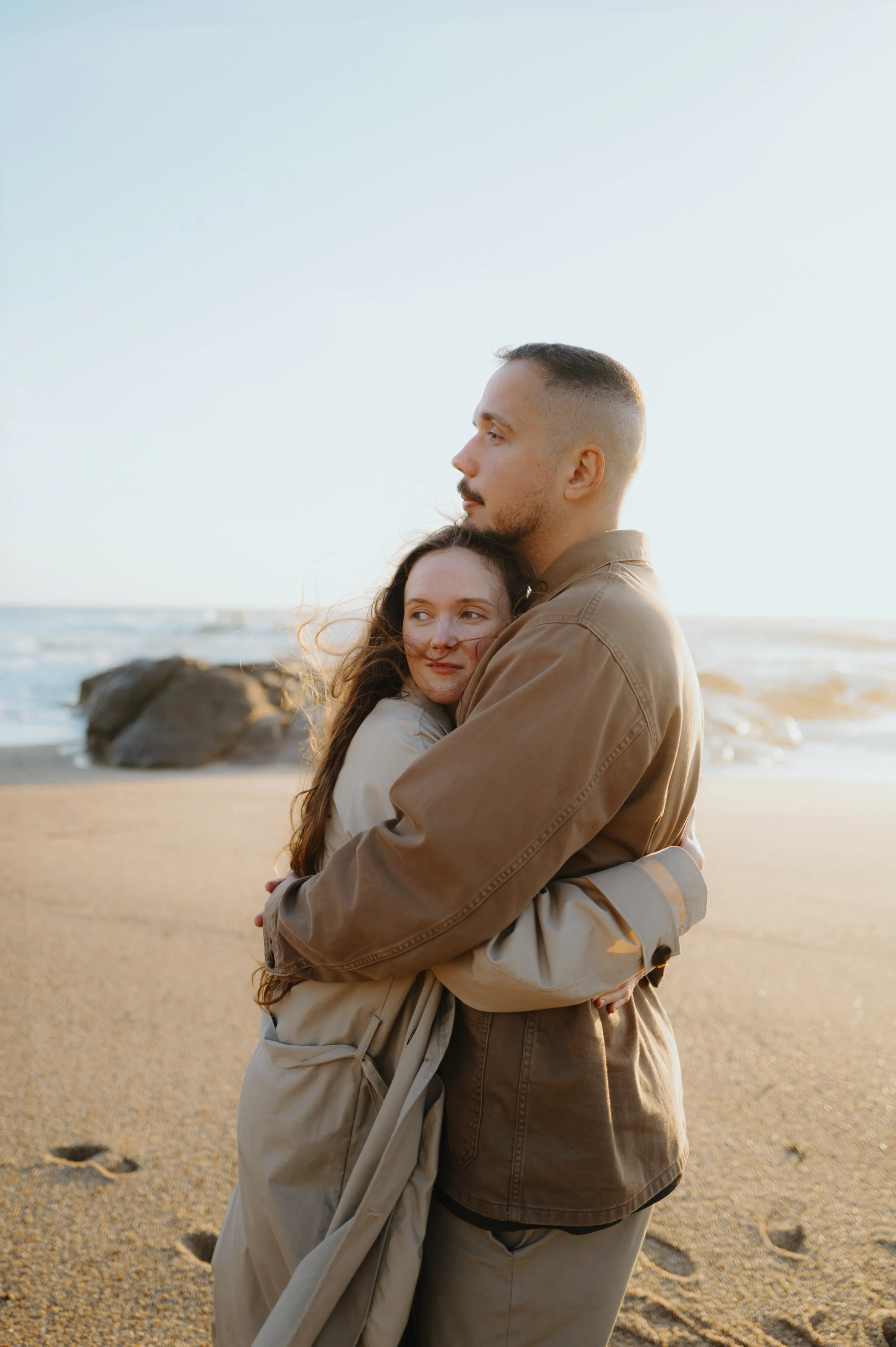 Couple embracing during a soft light seaside portrait on a sandy beach with rocks behind them.