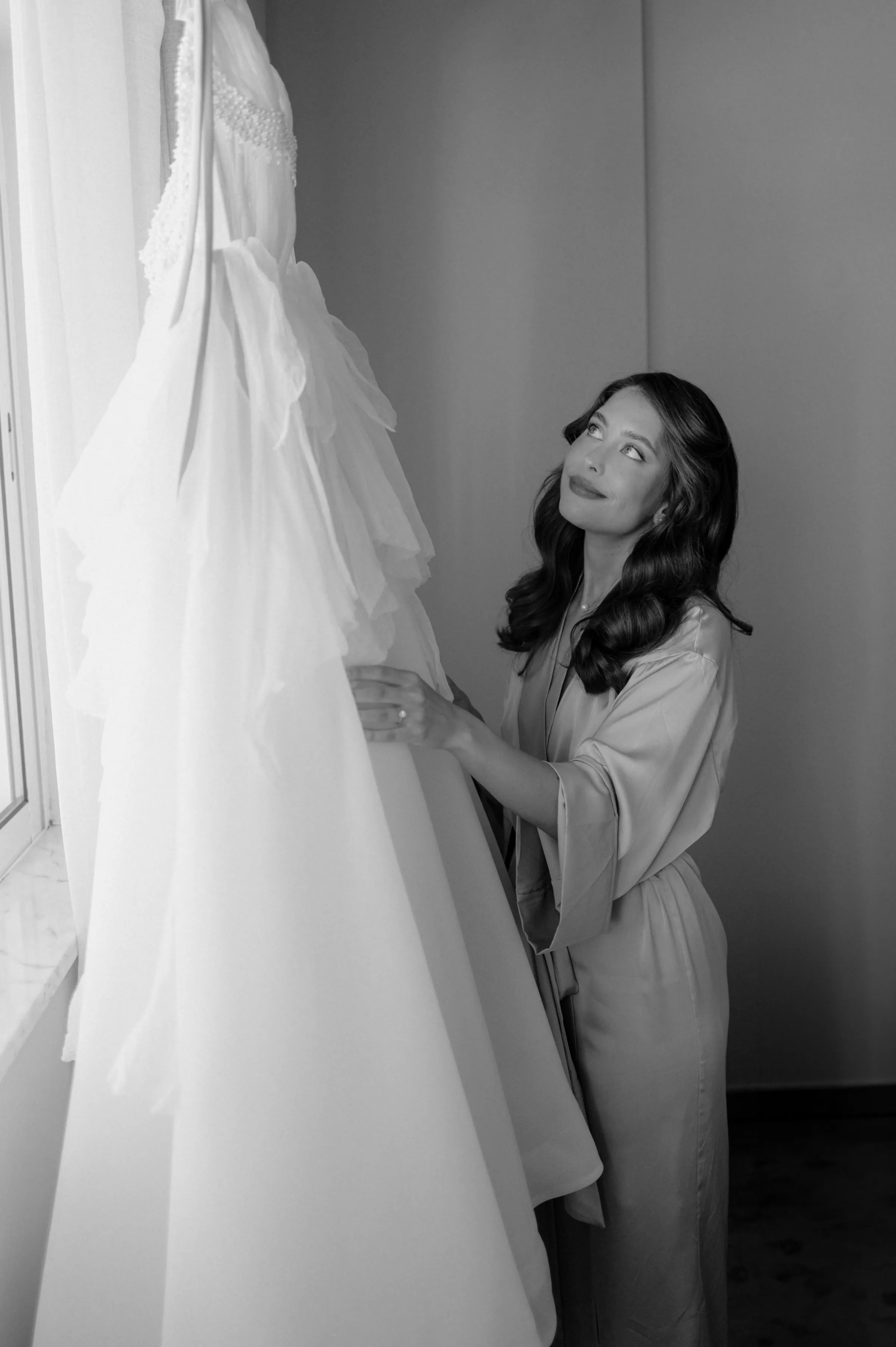 Black-and-white portrait of a woman beside a hanging gown in soft window light.