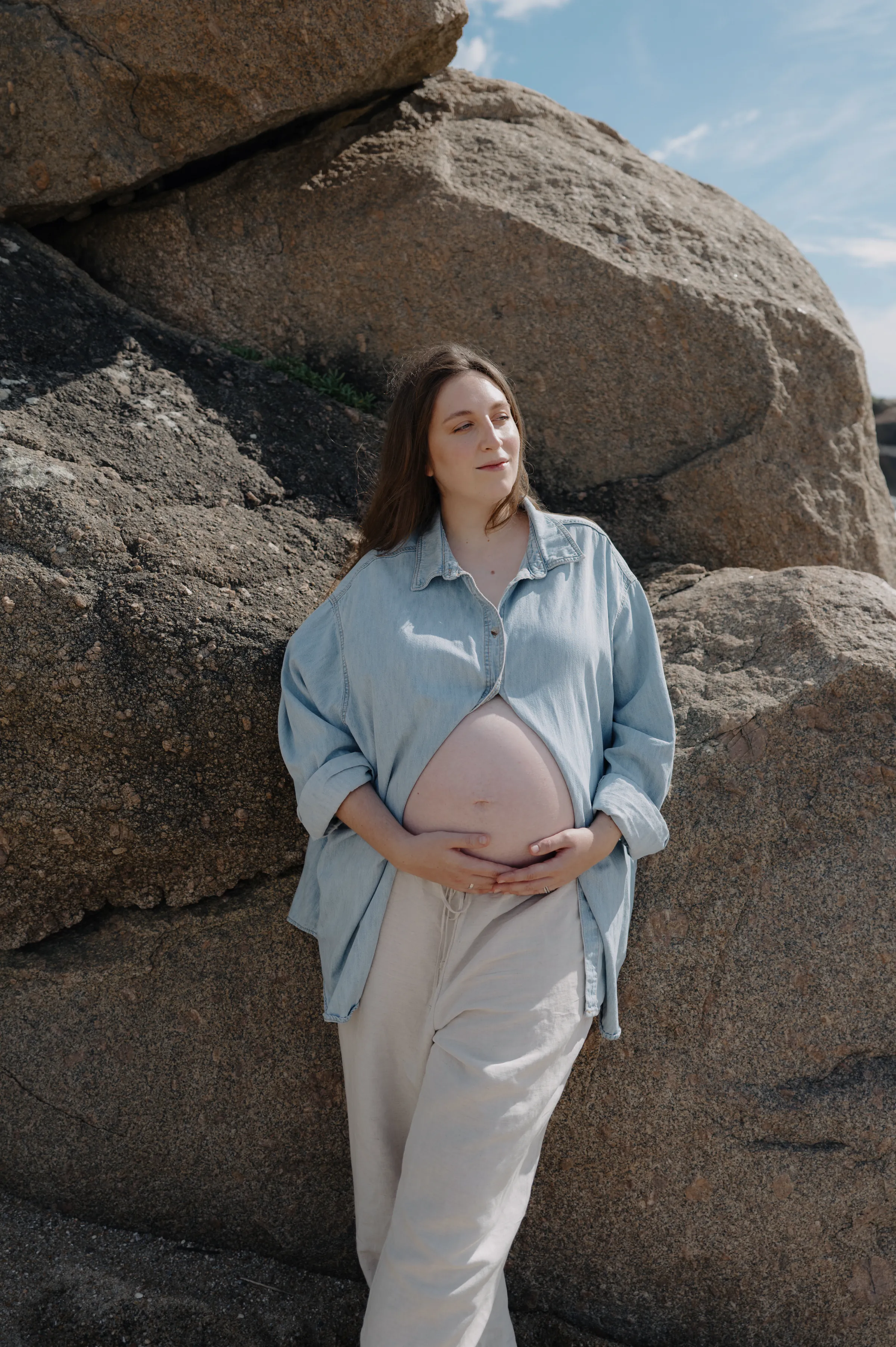 Outdoor portrait beside large rocks, person in pale layers resting hands on a rounded belly.