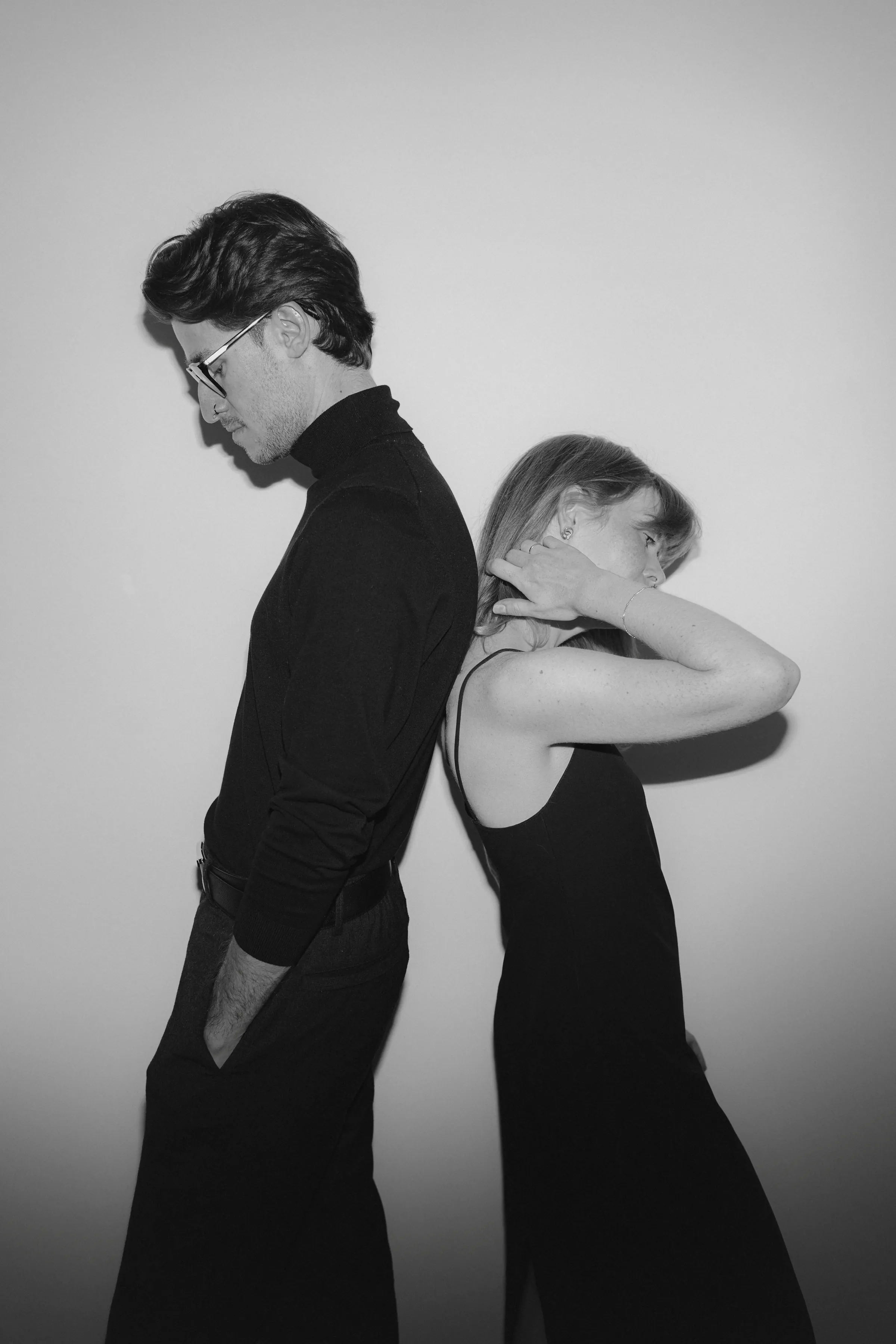 Black-and-white couple portrait standing back-to-back against a plain wall in direct light.