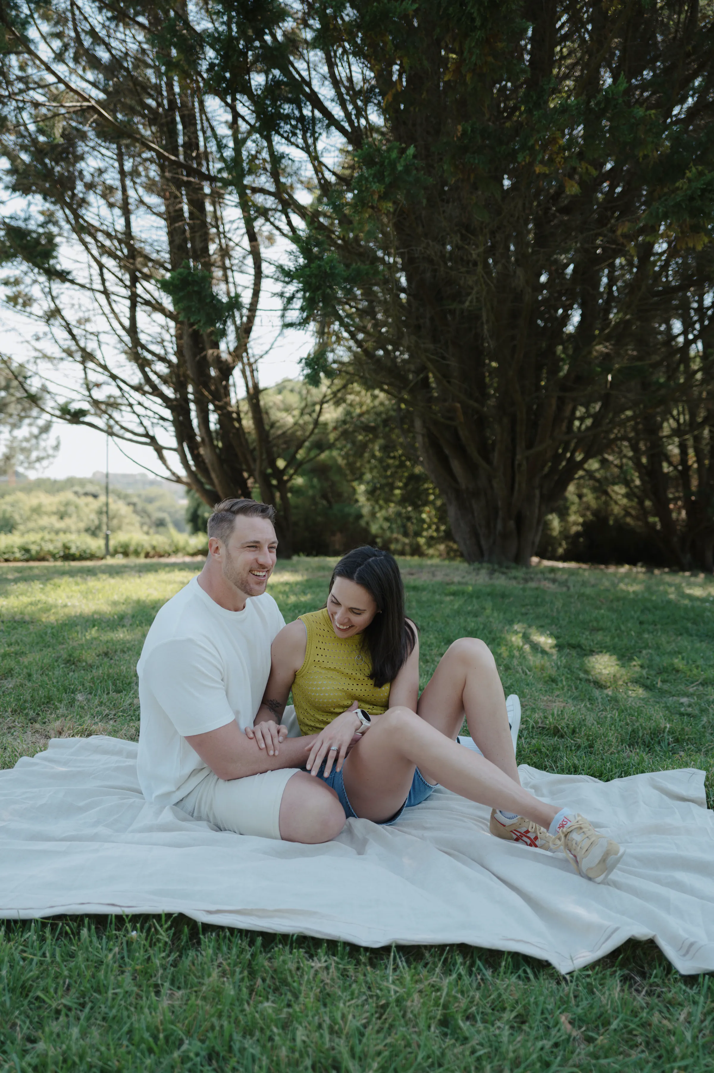 Couple sitting on a blanket during a relaxed outdoor session in a grassy park under tall trees.