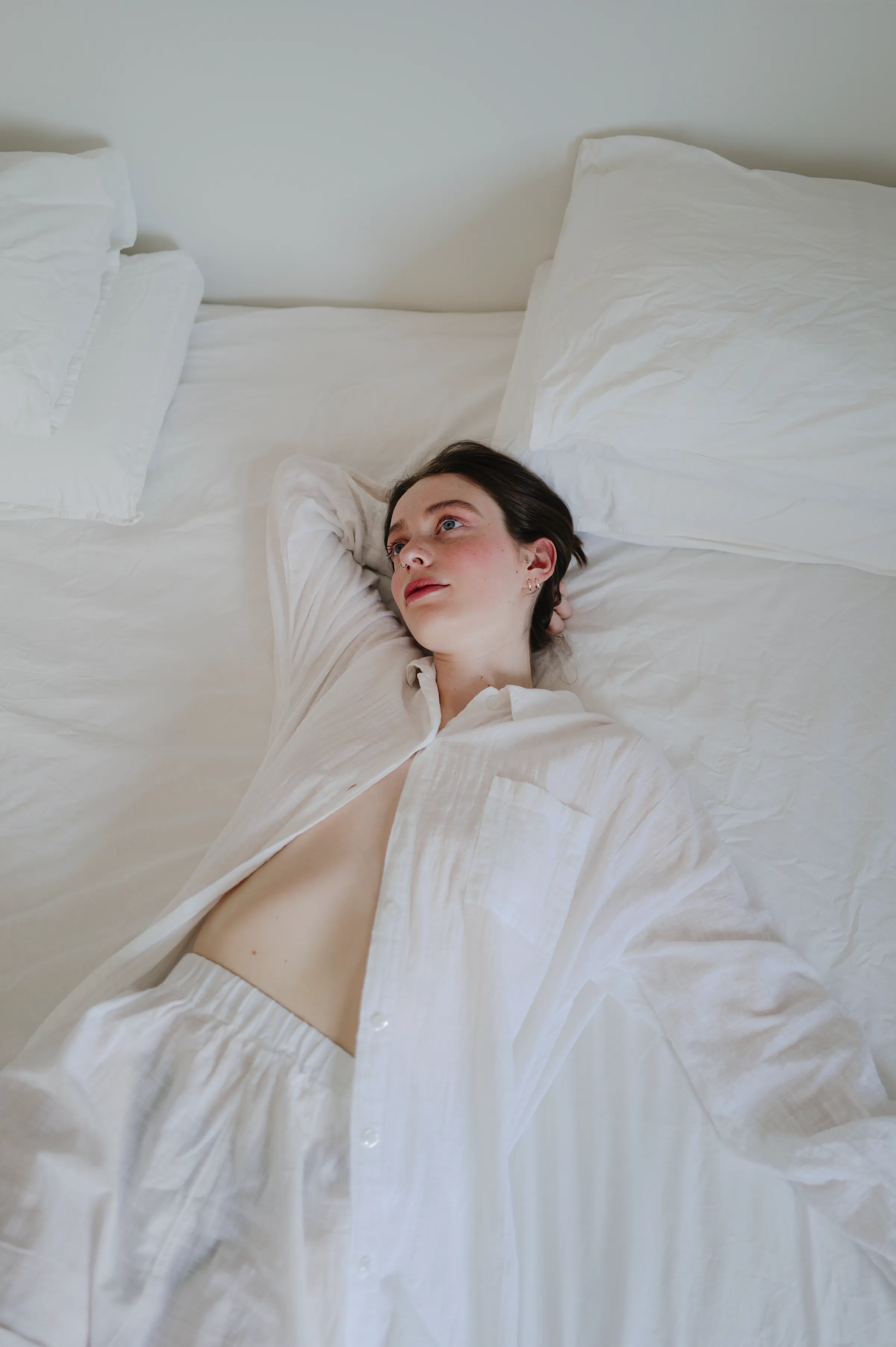 Overhead portrait of a person reclining on white bedding in soft indoor light.