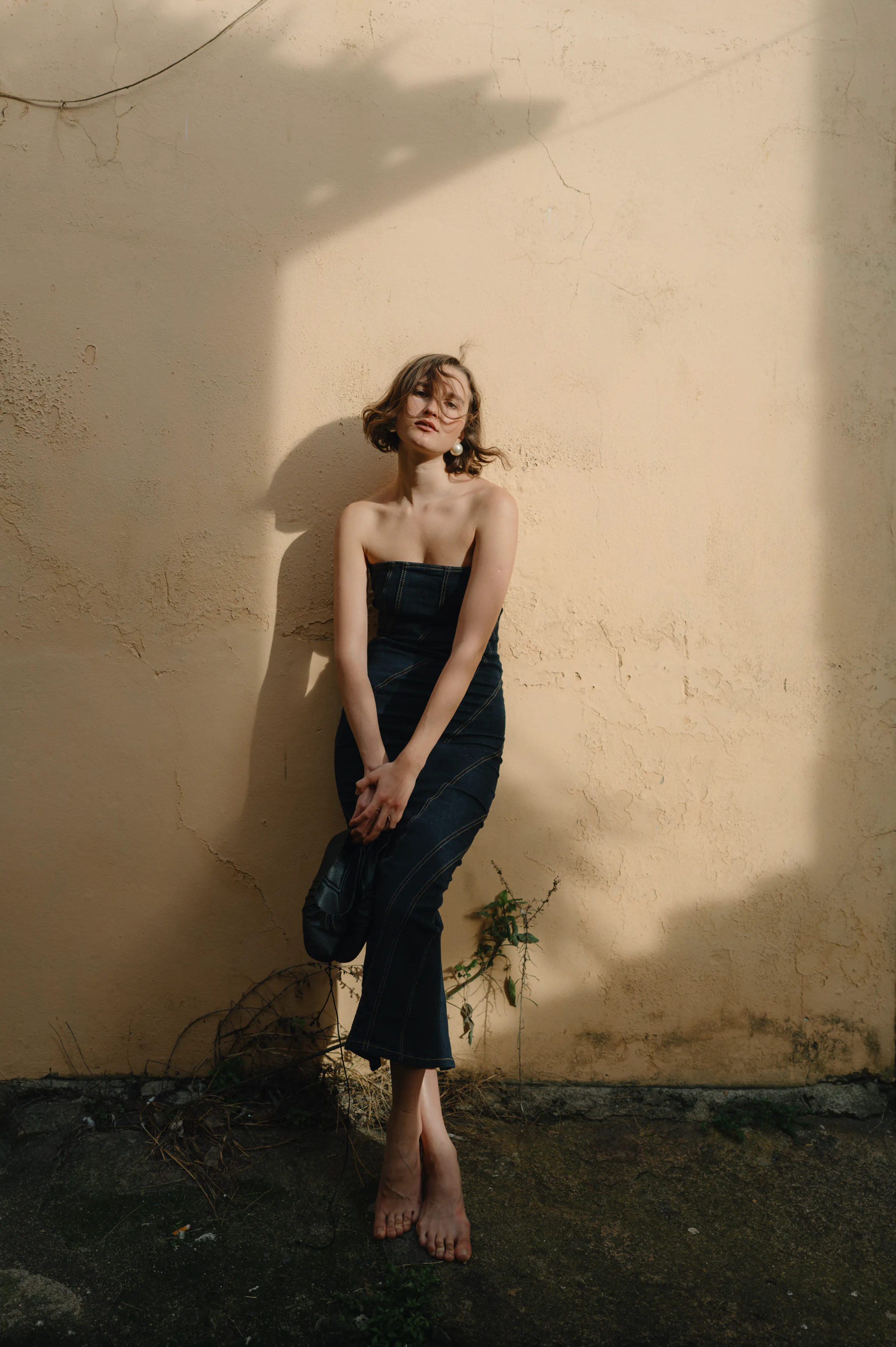Outdoor portrait of a barefoot person in a dark strapless dress against a sunlit textured wall.