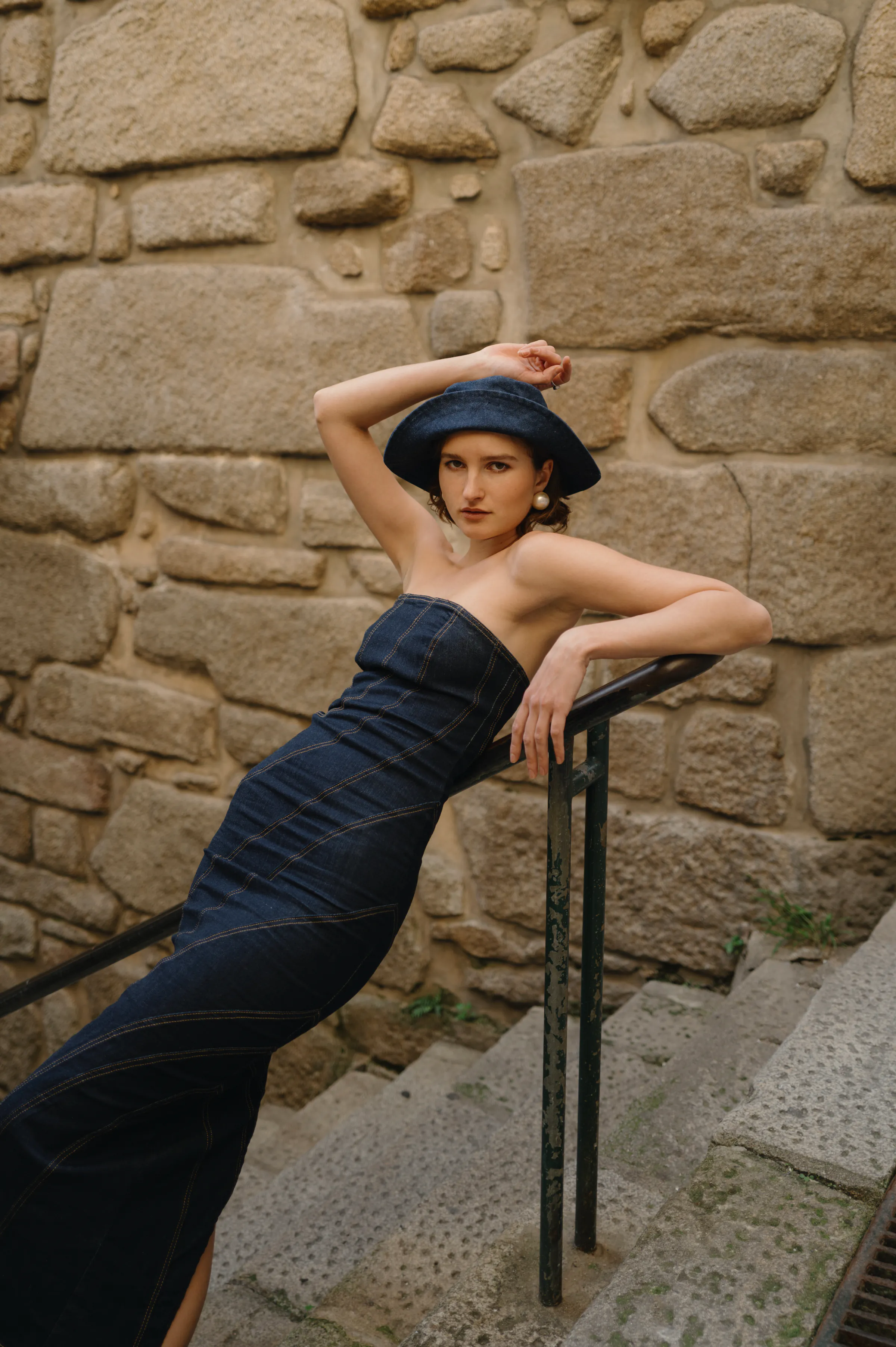 Fashion portrait of an adult in a dark dress and hat posing on stone steps by a textured wall.