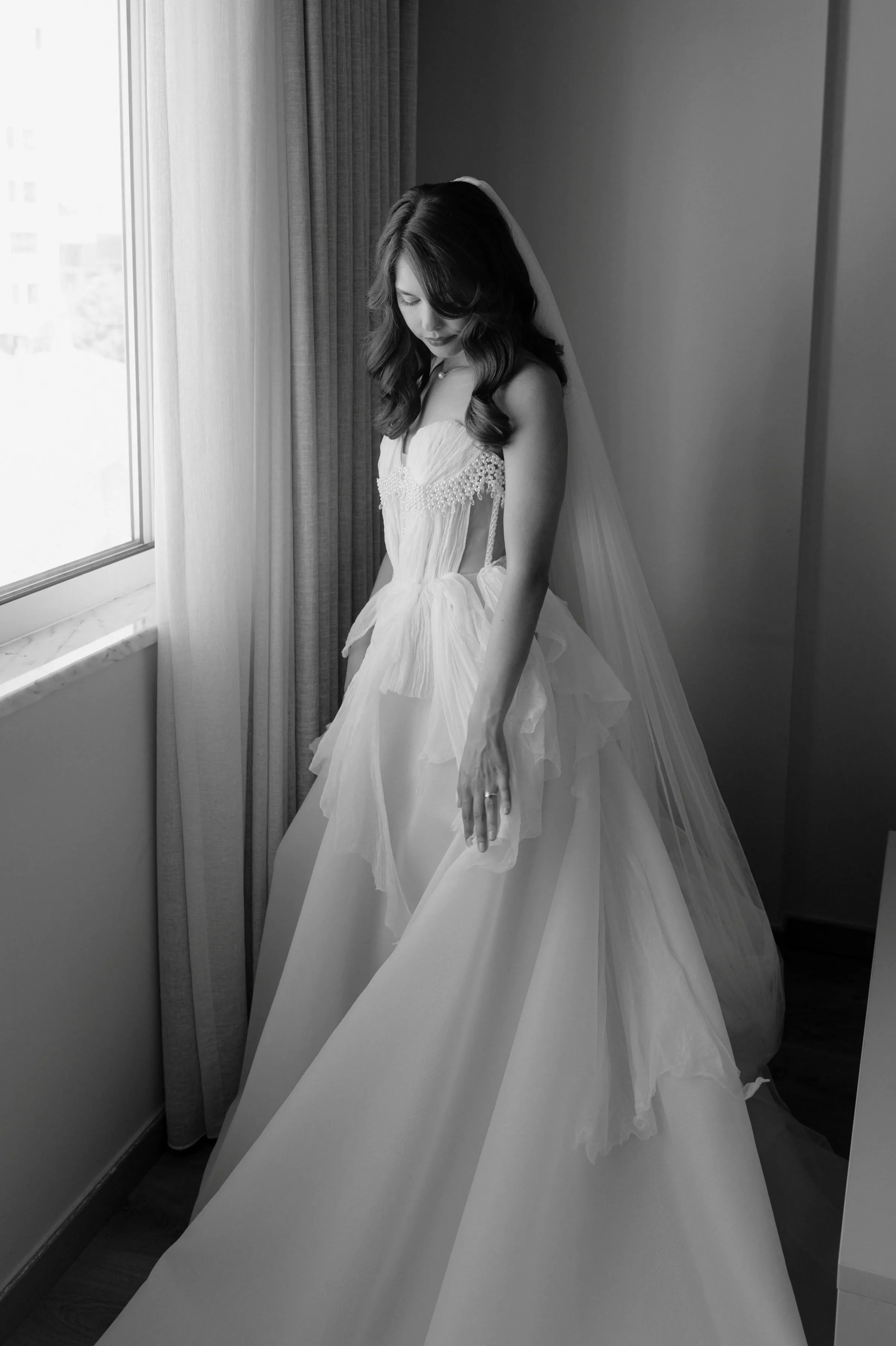 Black-and-white bridal portrait beside a window, with soft light on a wedding gown and veil.