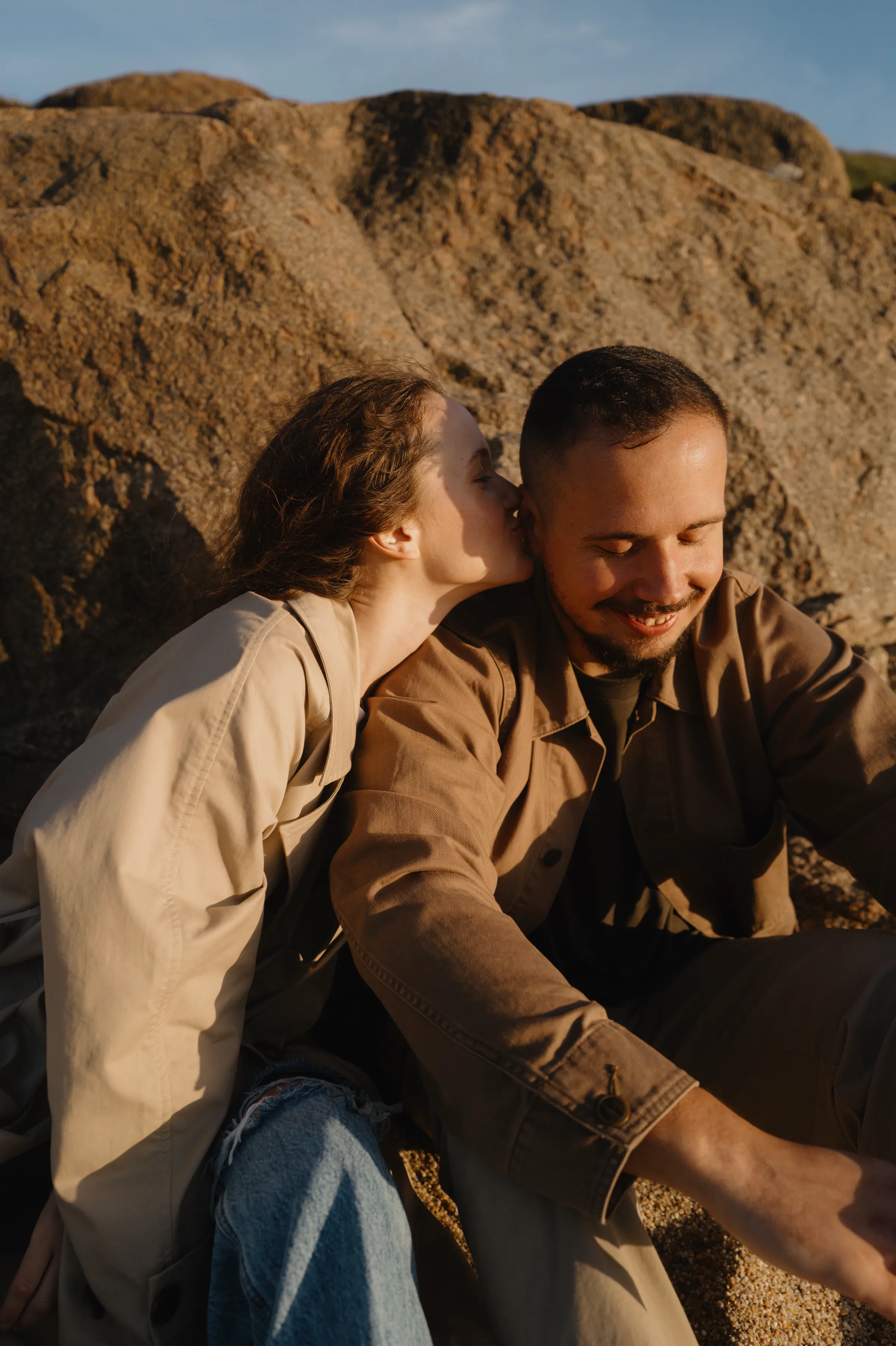 Couple seated by sunlit rocks during an outdoor session, one kissing the other’s cheek in soft light.