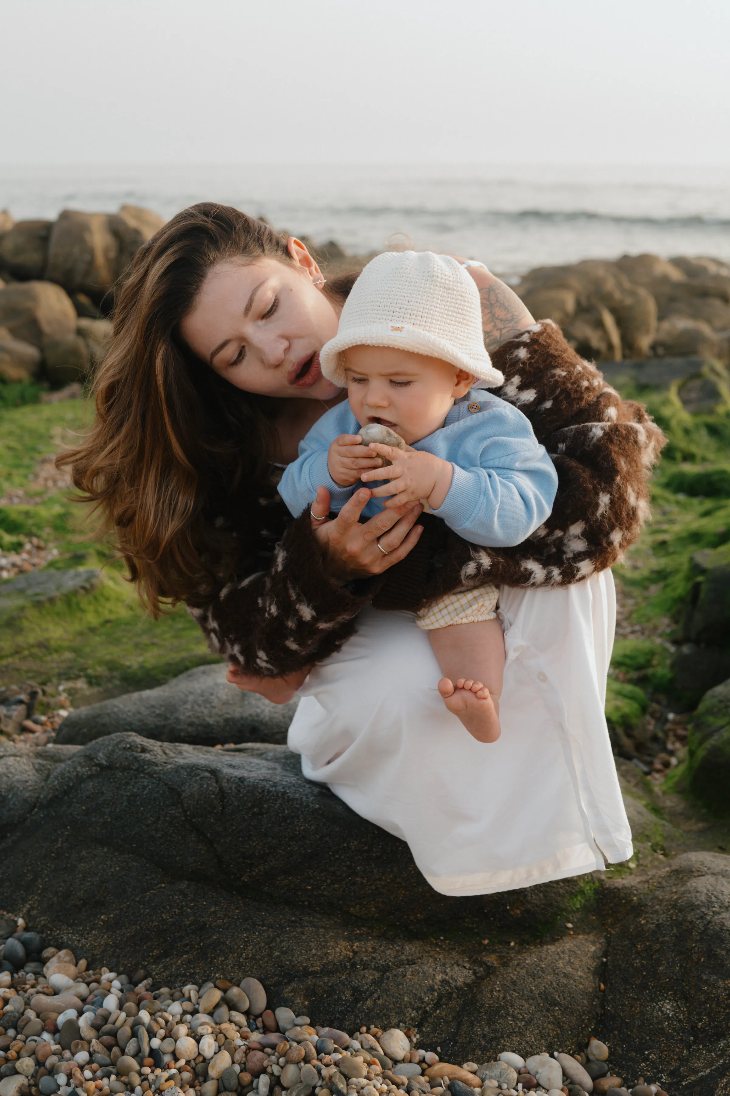 Adult holding a baby during a seaside outdoor session on coastal rocks in soft light.
