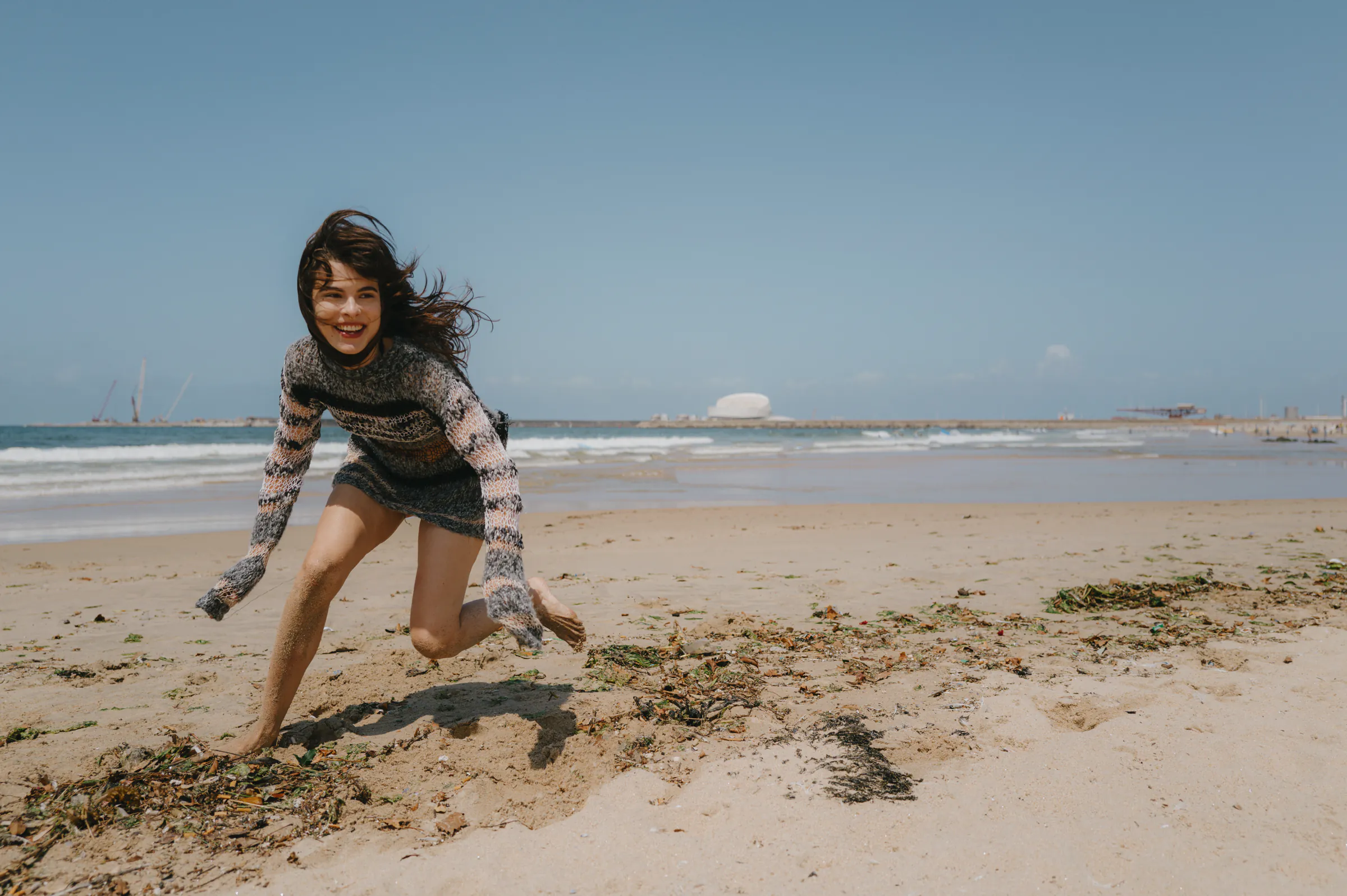 Seaside portrait of a woman crouching on sandy beach, hair windblown under clear sky.