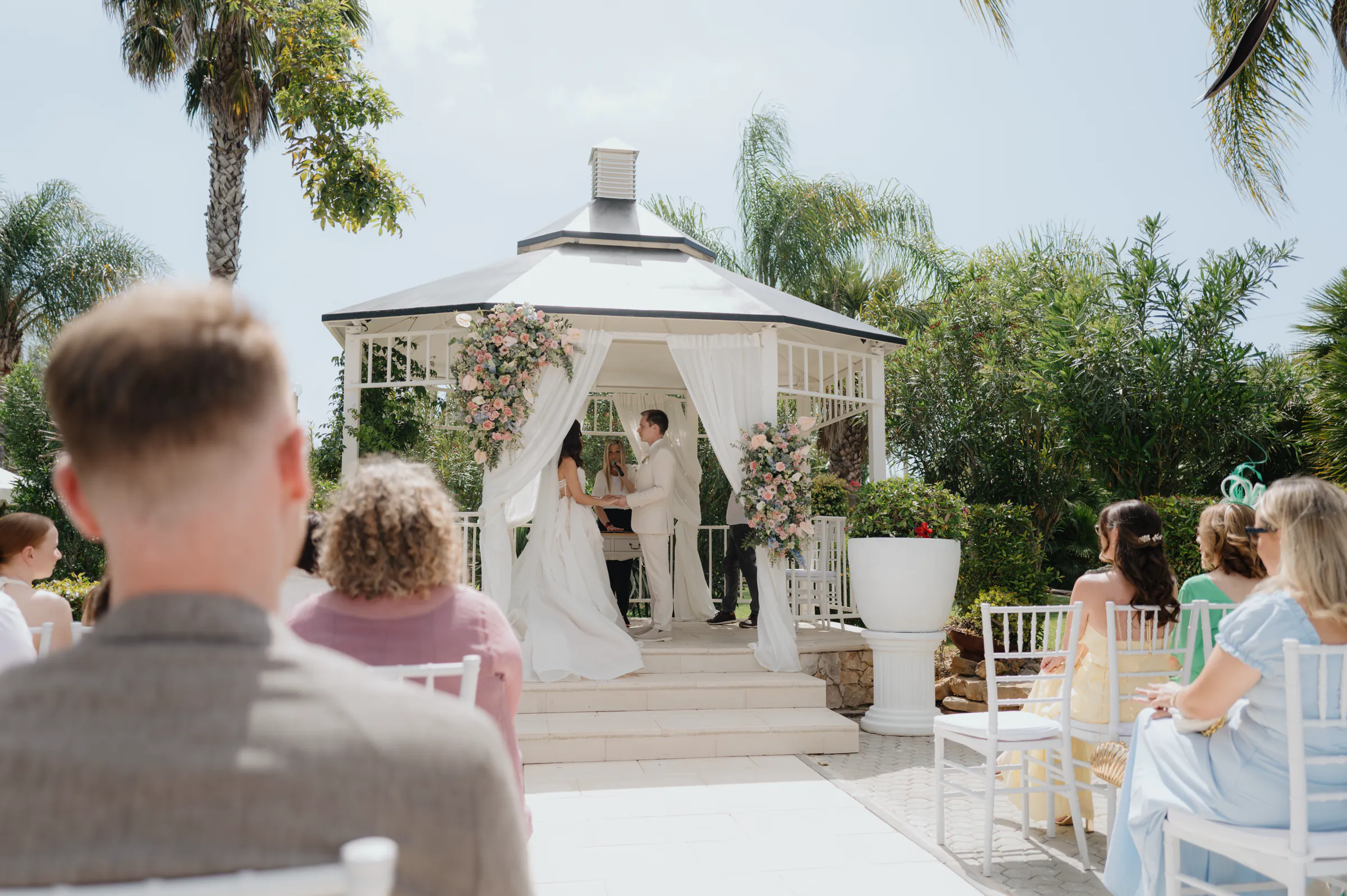 Couple standing in a white gazebo during an outdoor ceremony with seated guests and palm trees.