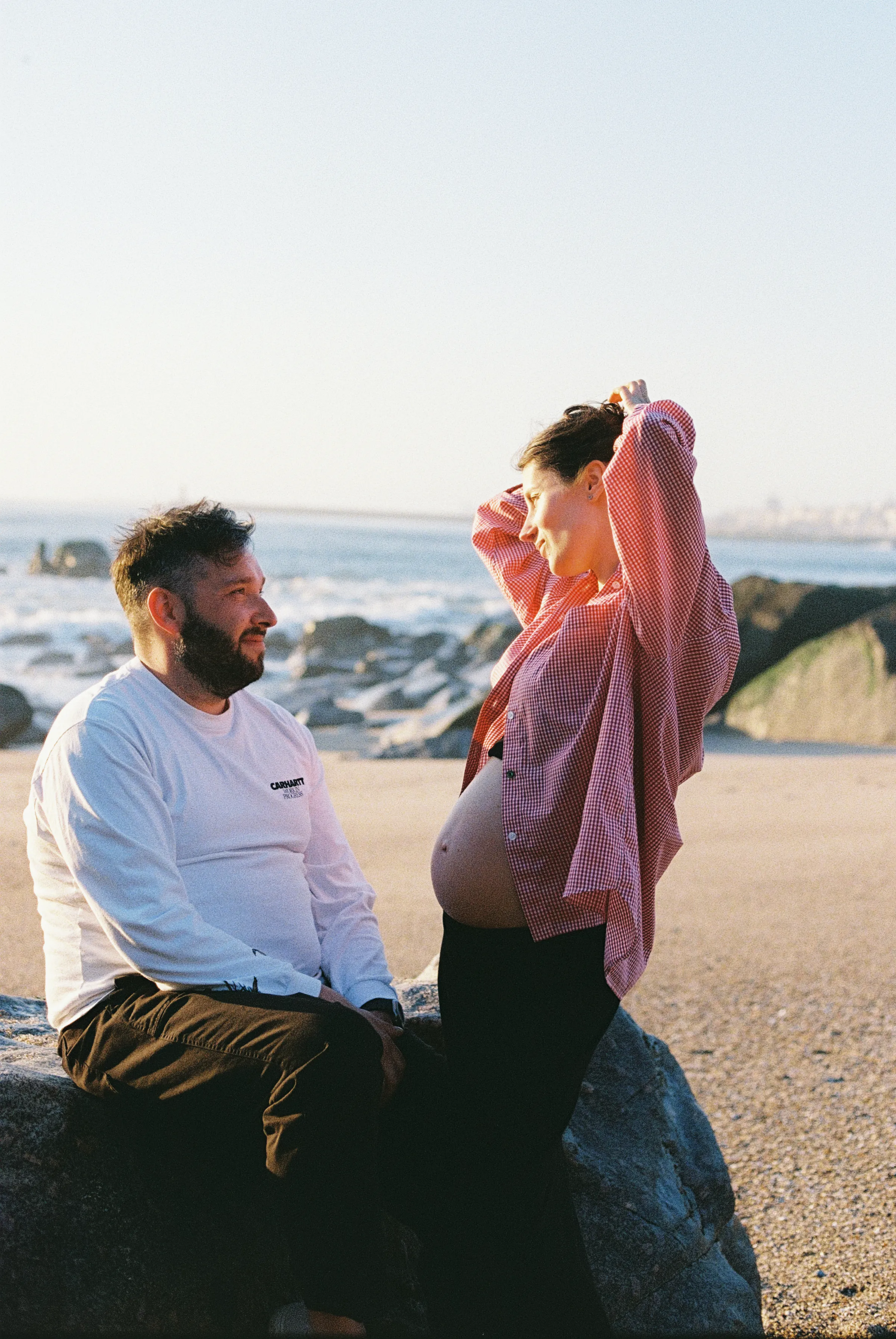 Couple during a seaside outdoor session, one standing by rocks as the other sits in soft light.
