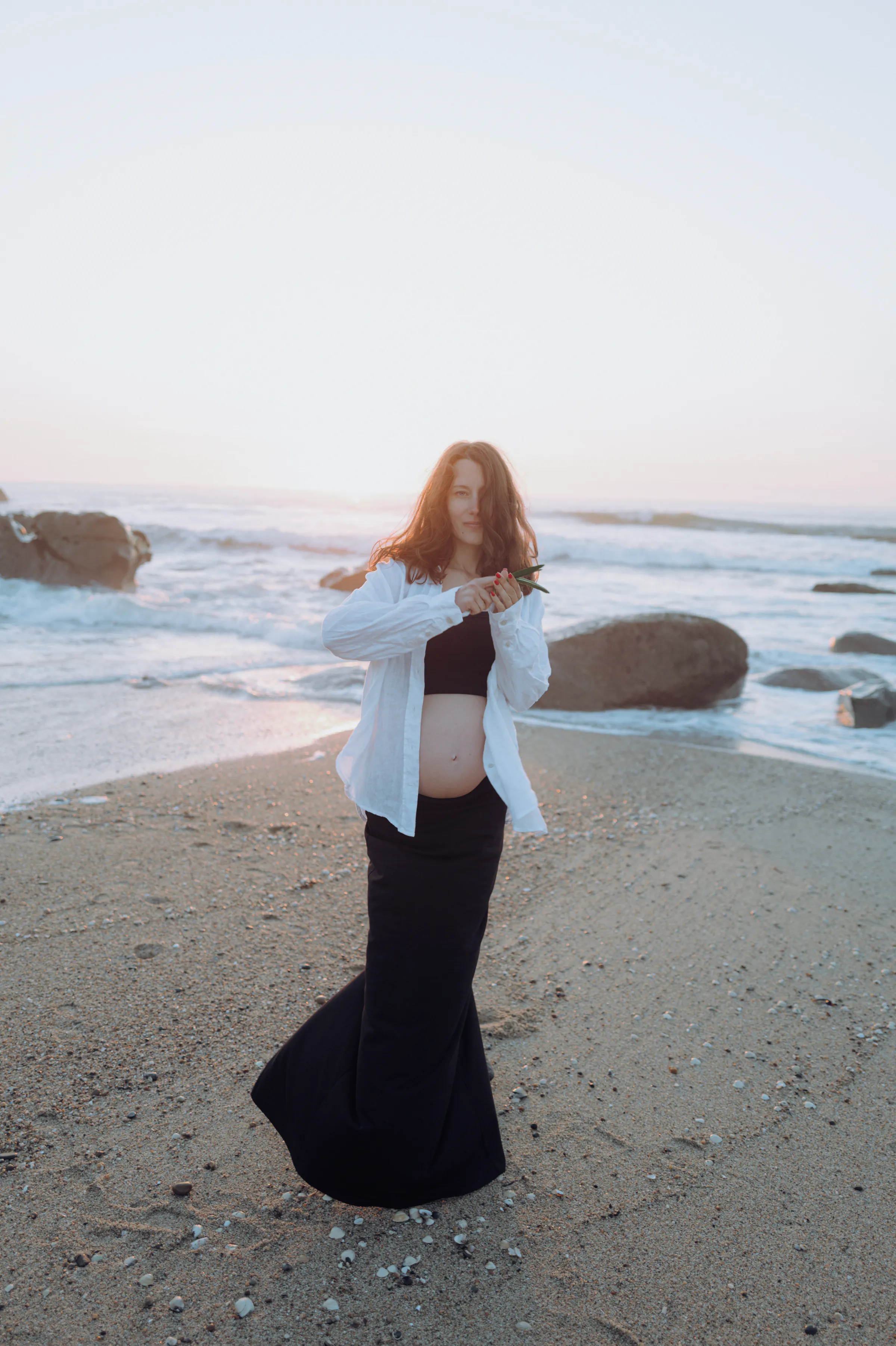 Seaside portrait of an adult standing on a sandy beach in soft light, with waves and rocks behind.
