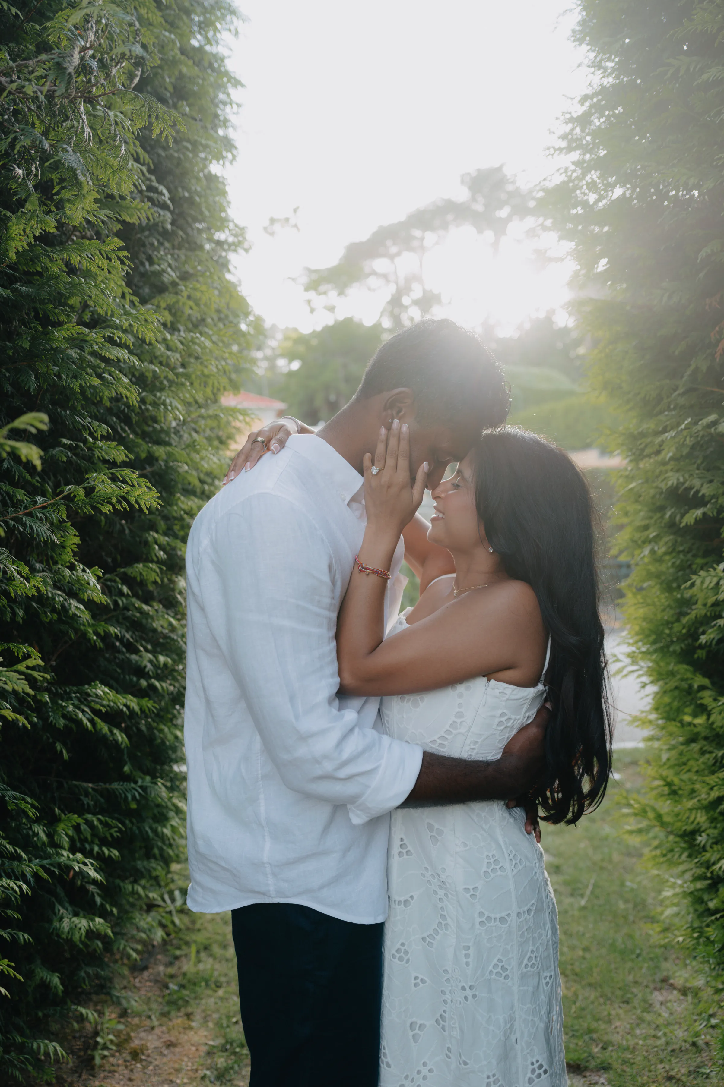 Couple in a close outdoor portrait on a hedge-lined garden path with soft backlight.