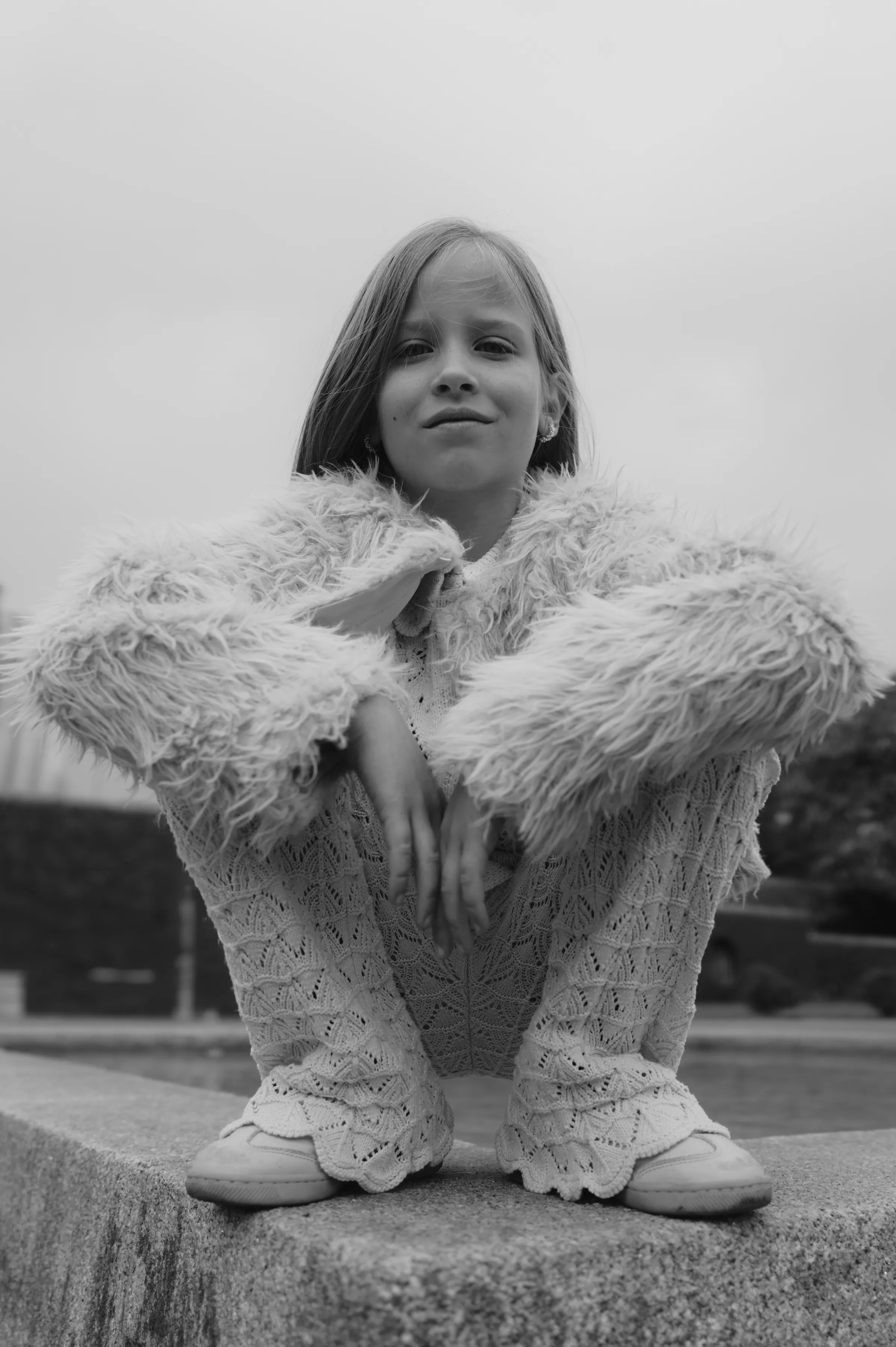 Black-and-white outdoor portrait of a child crouching on a stone ledge in a fluffy coat.