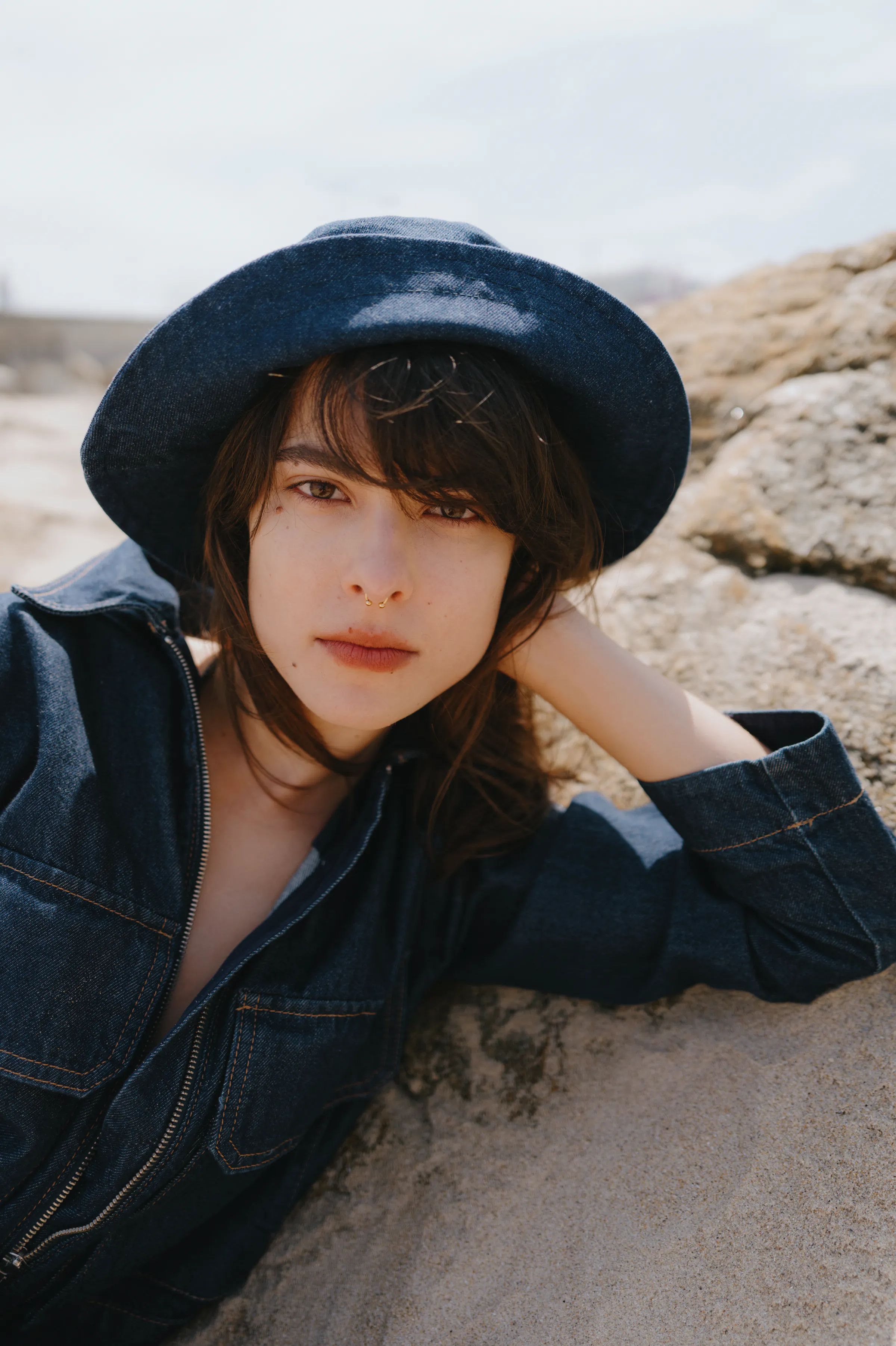 Close portrait of a person in a dark hat and denim jacket reclining on seaside rocks in soft light.