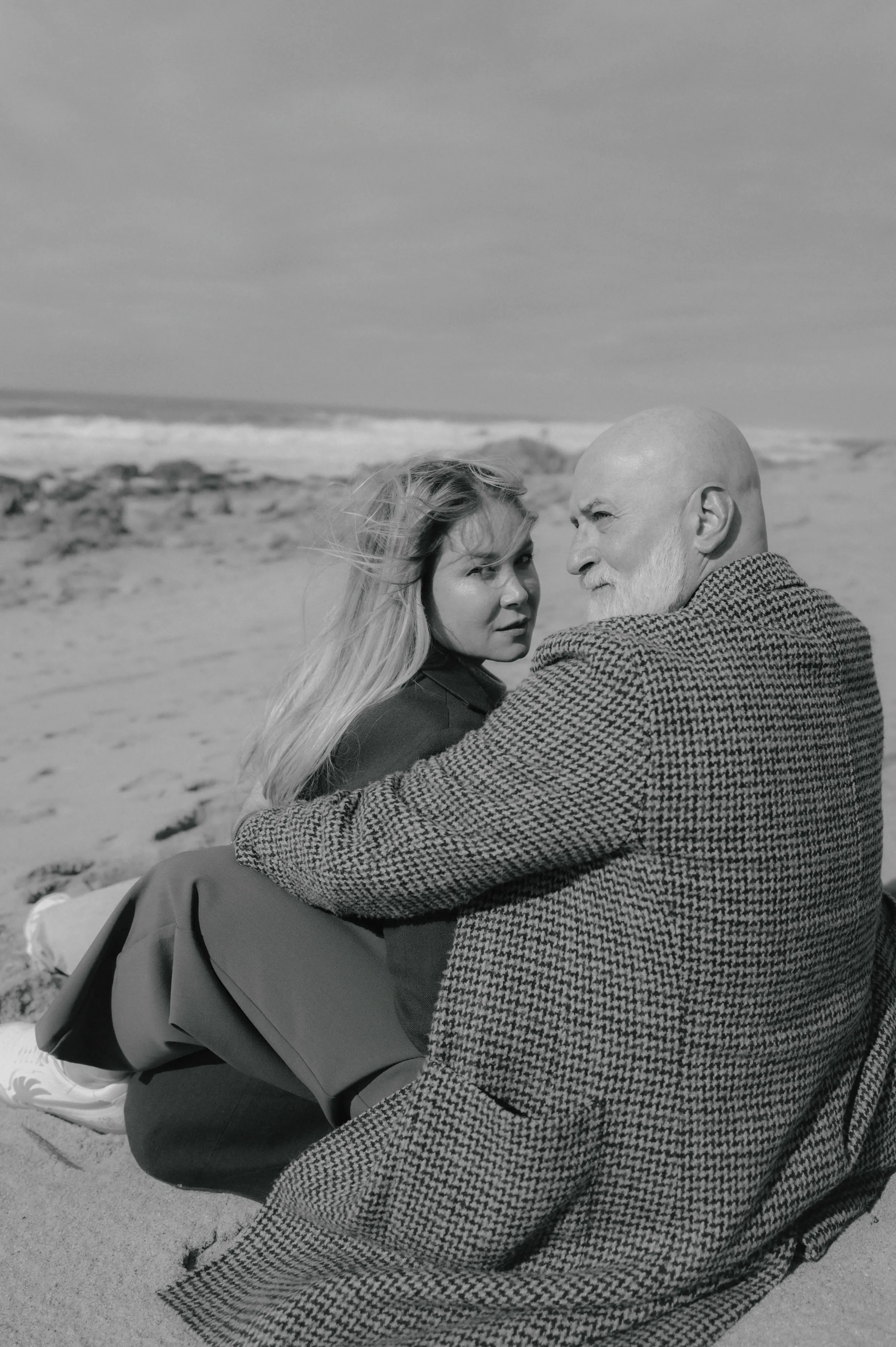 Black-and-white seaside portrait of a couple seated close together on the sand in soft light.