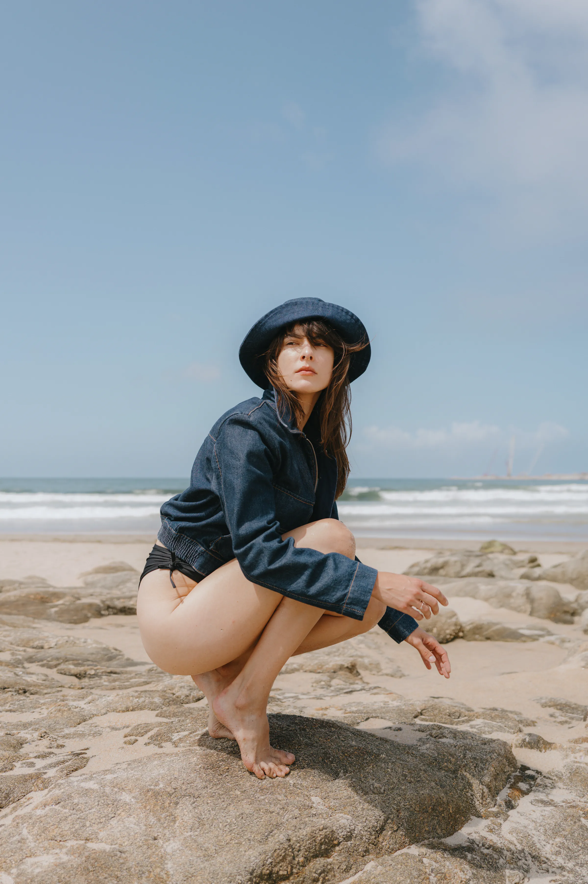 Seaside portrait of an adult crouching on rocks in a dark hat and denim shirt.