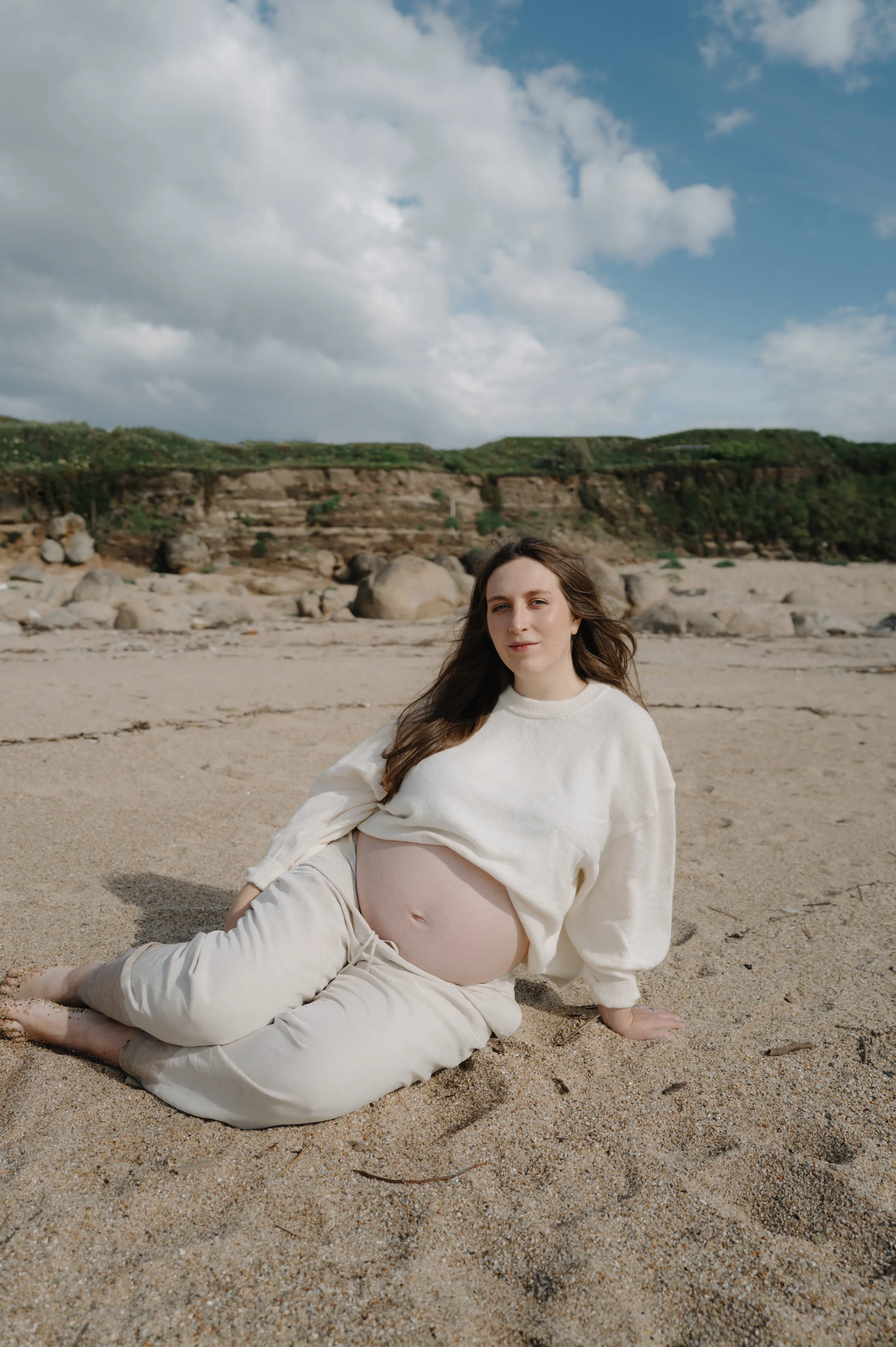 Seaside portrait of a person in white reclining on sandy sand beneath rocky cliffs and soft cloudy light.