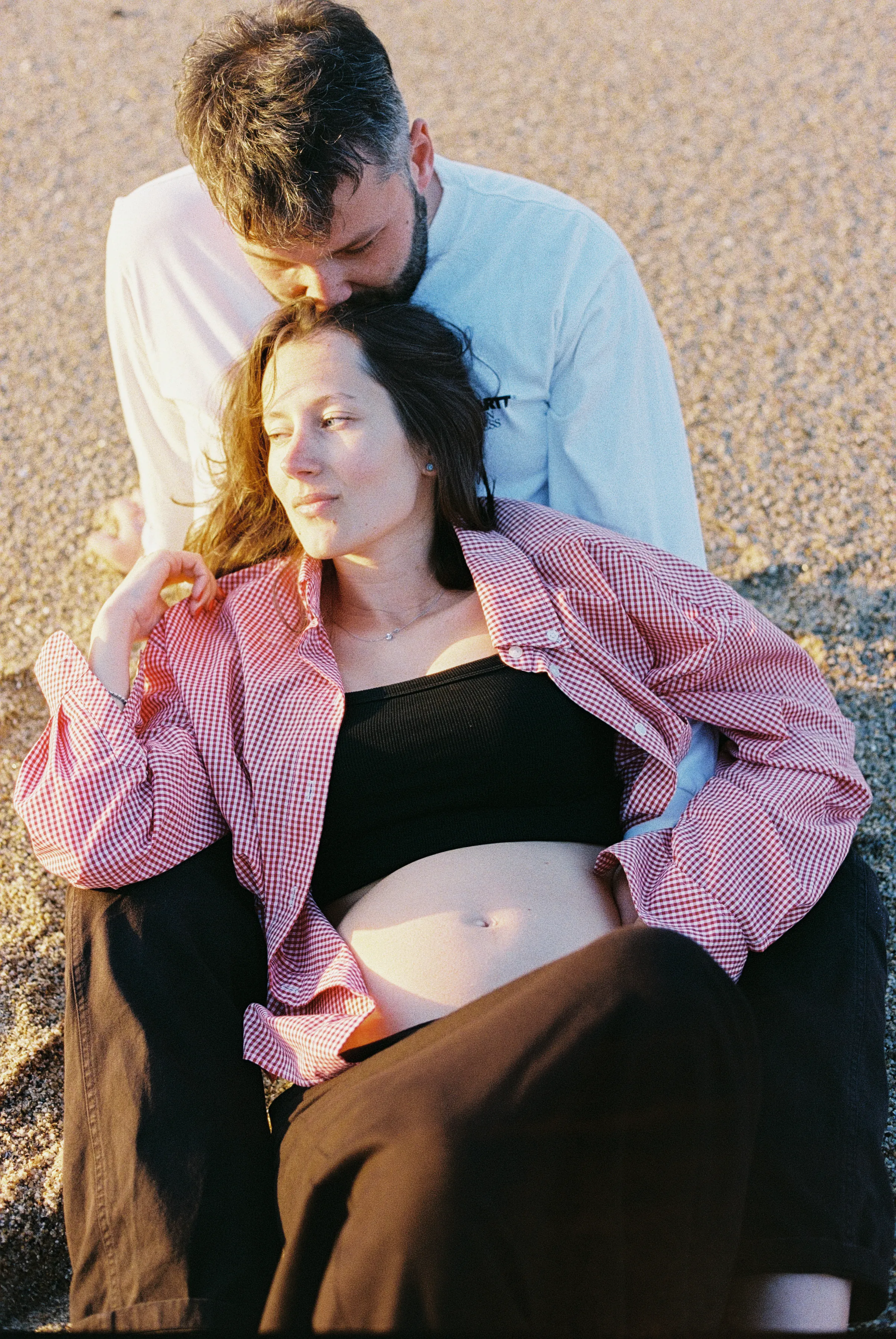 Pregnant couple reclines together on a pebbled beach during a soft light outdoor session.