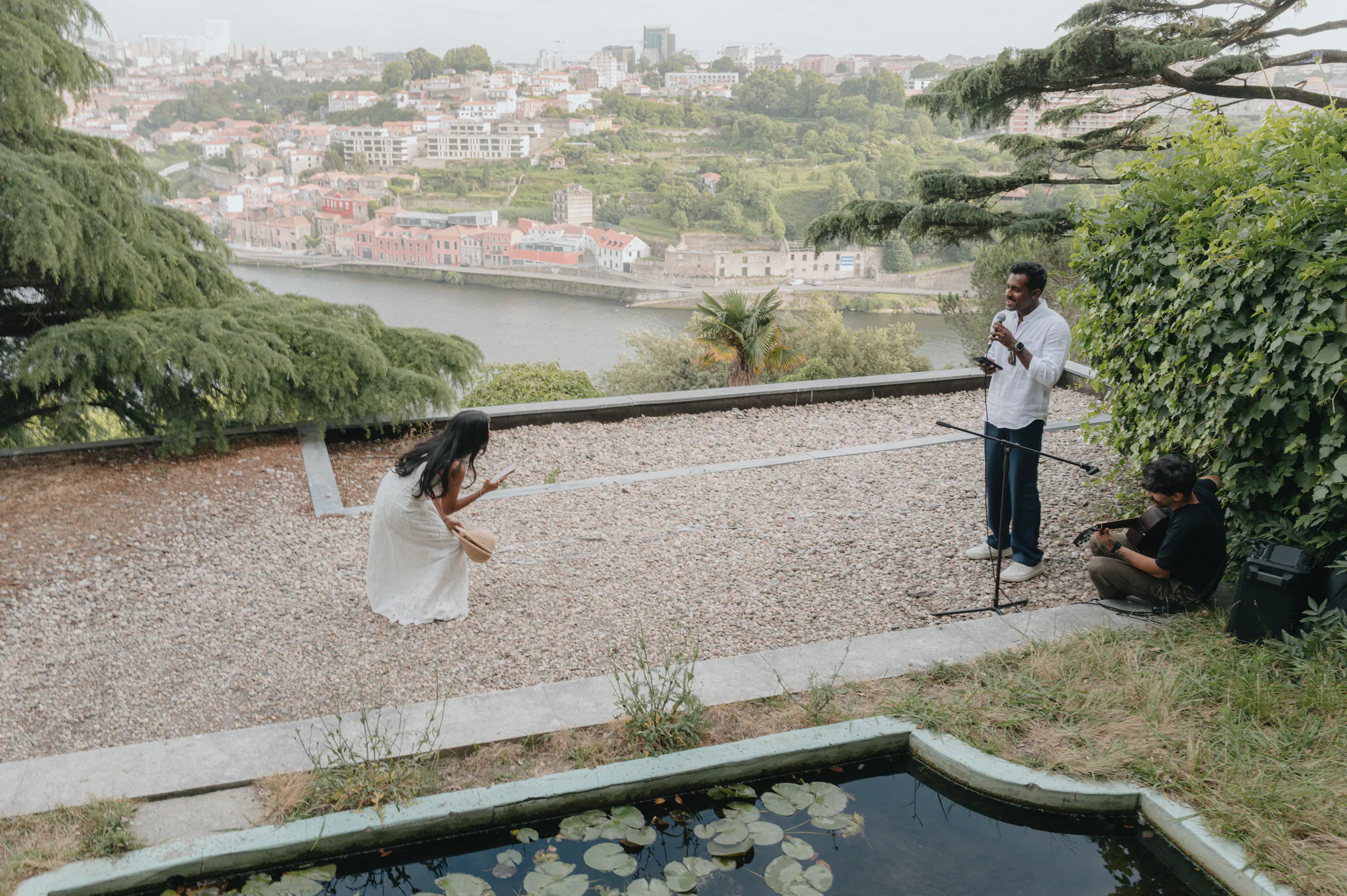 Outdoor session on a garden terrace overlooking a river city, with a woman in white and camera crew nearby.