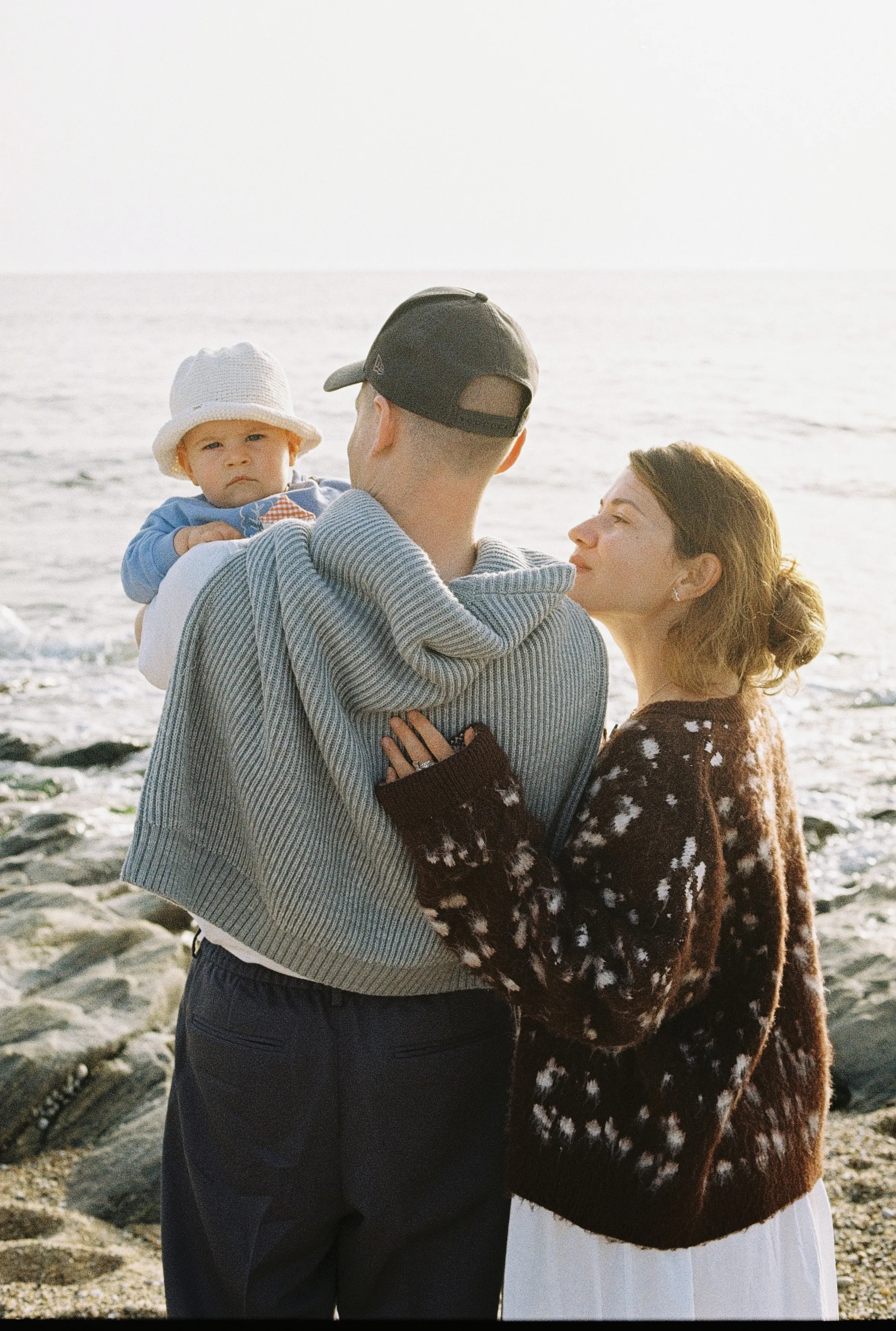 Family portrait on rocky seaside, with a baby held close between two adults in soft light.