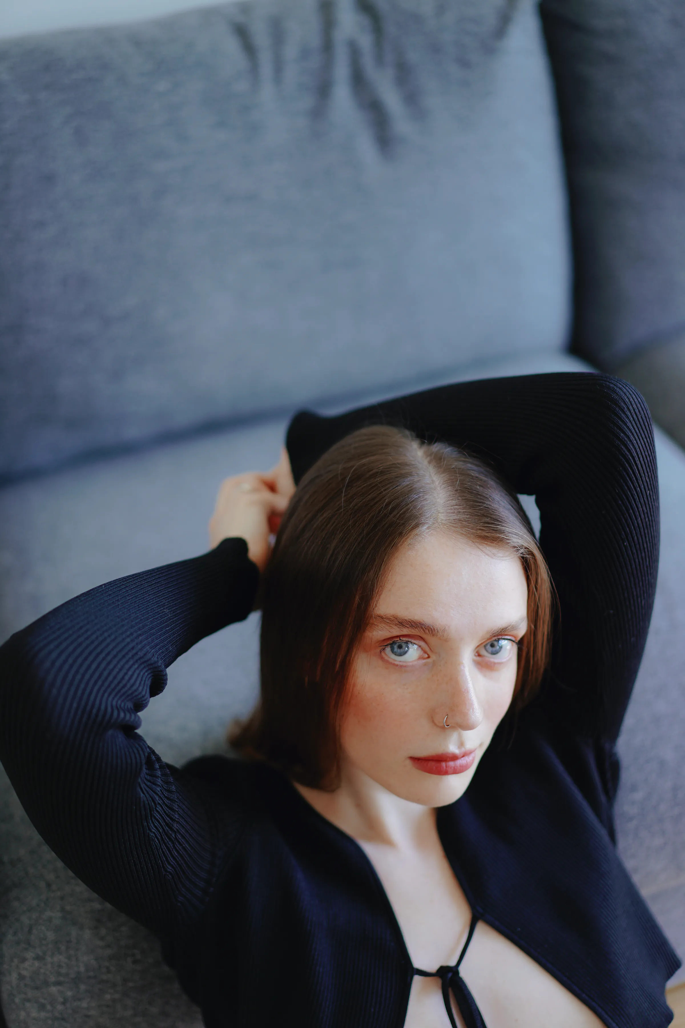Close portrait of a person reclining on a gray sofa, arms raised, in soft indoor light.