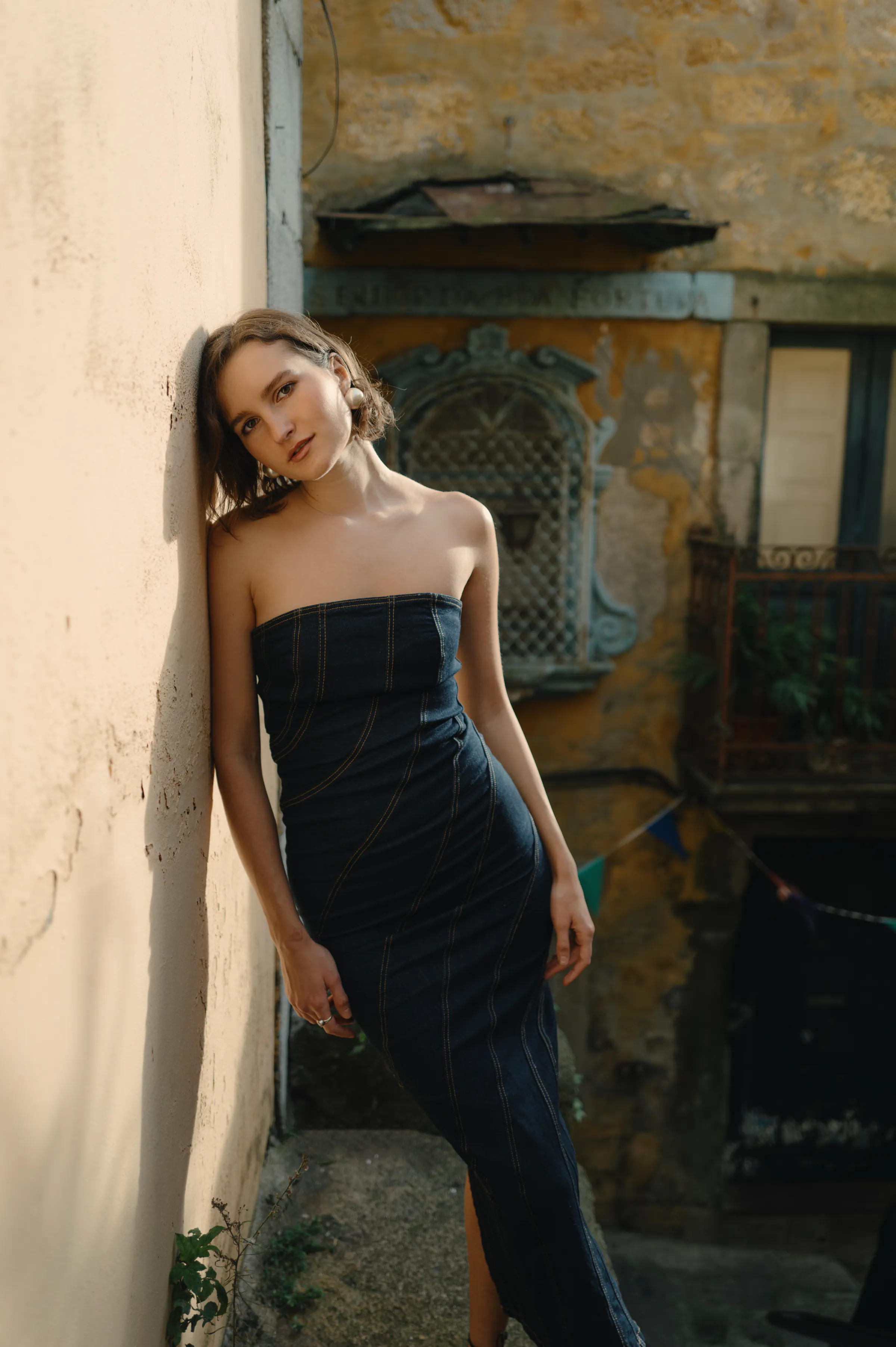 Outdoor portrait of a woman leaning against a sunlit wall in an old city courtyard.
