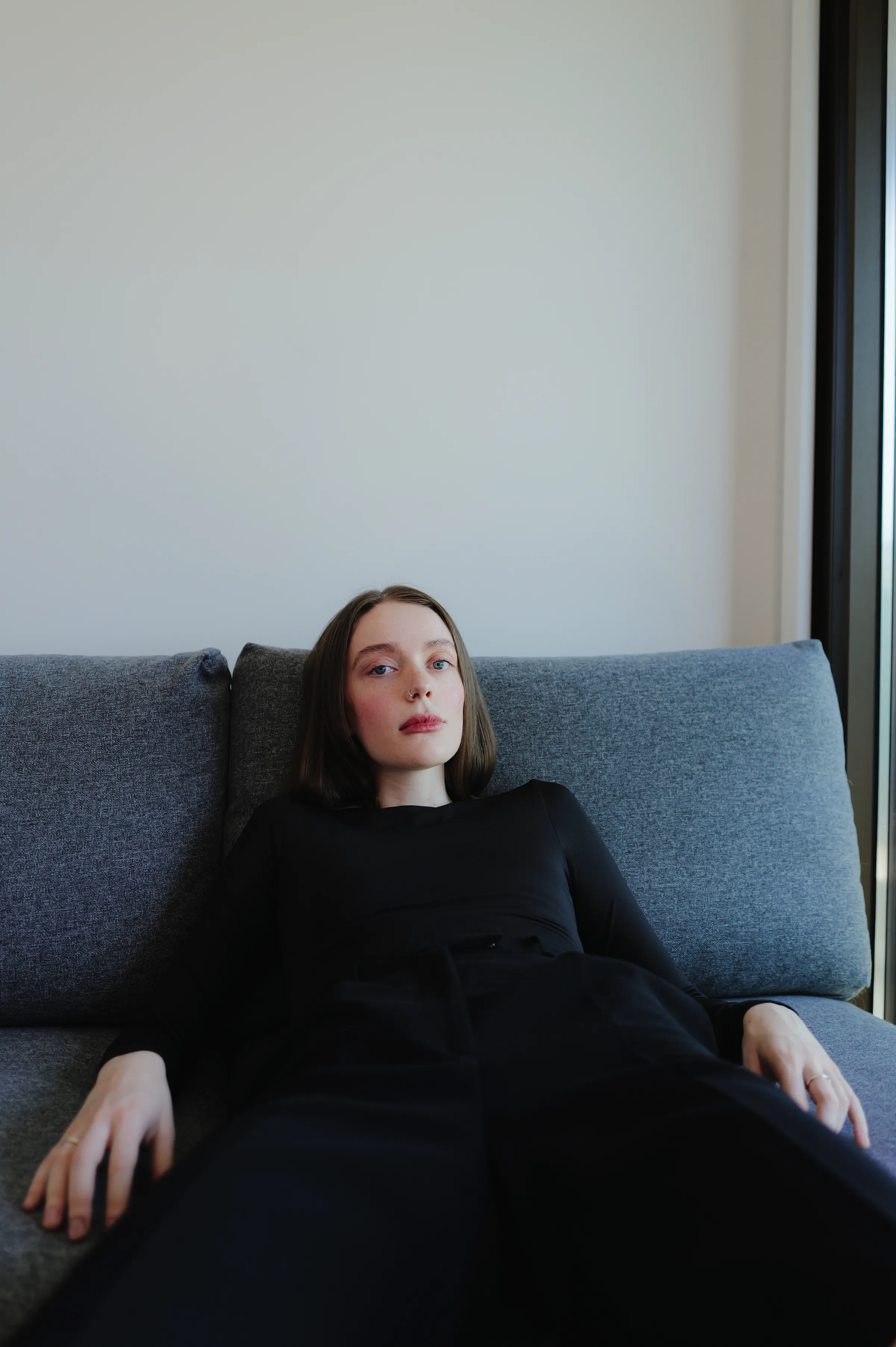 Indoor portrait of a person reclining on a gray sofa beside a window in soft light.