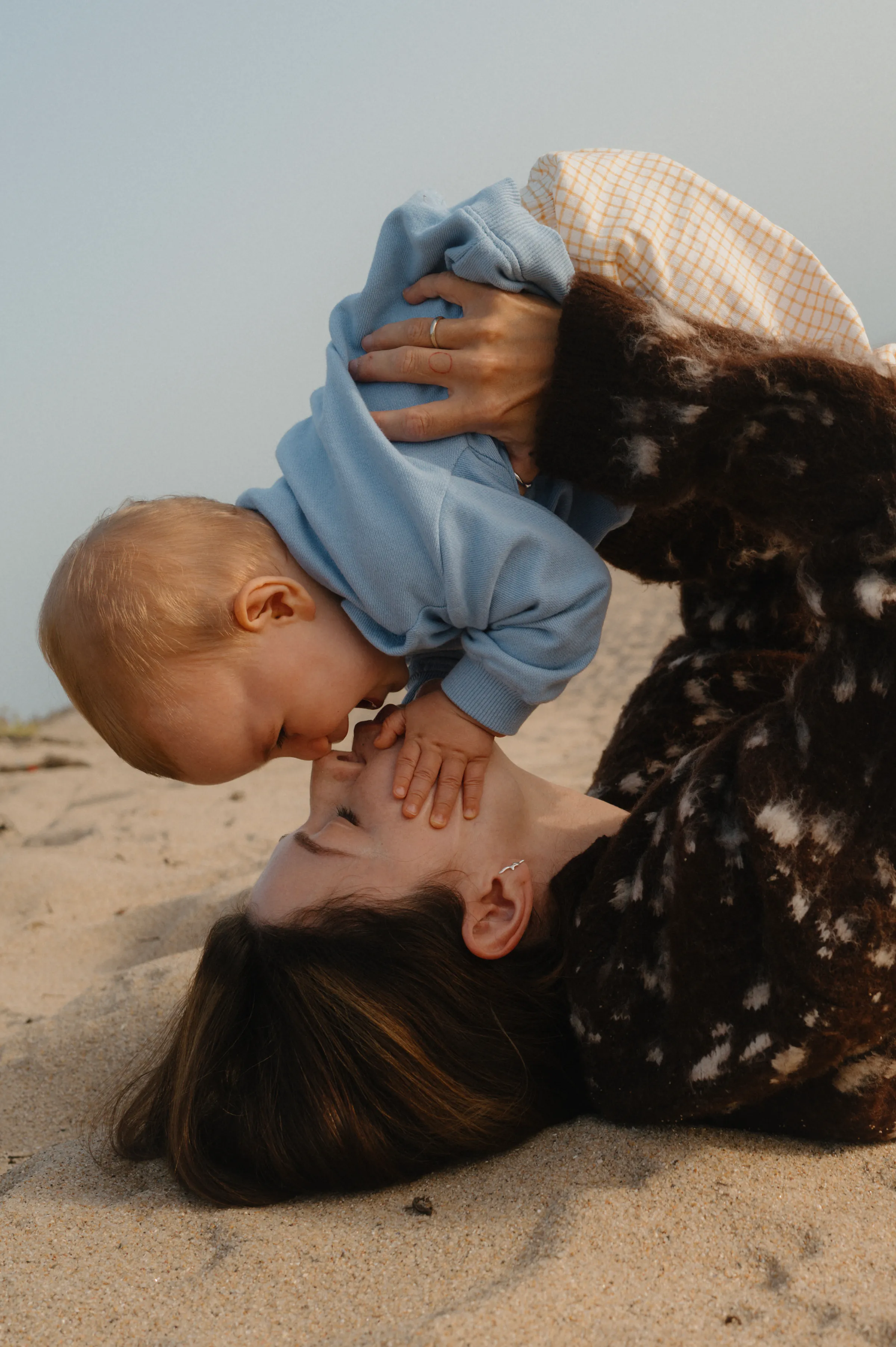 Baby leans toward an adult lying on sand during a soft light seaside family session.