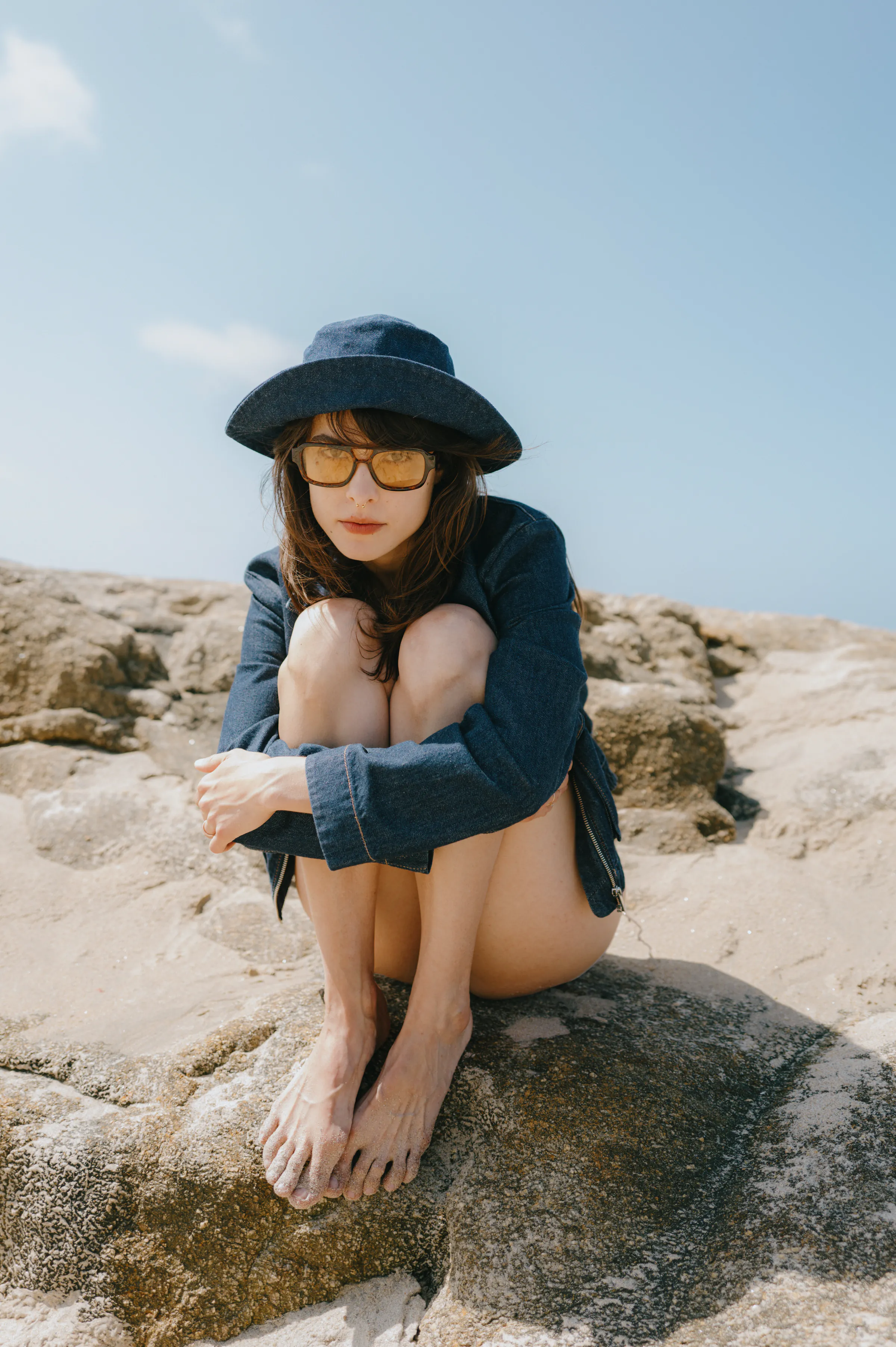 Outdoor portrait of an adult in a wide-brim hat and sunglasses, seated barefoot on sunlit rocks.